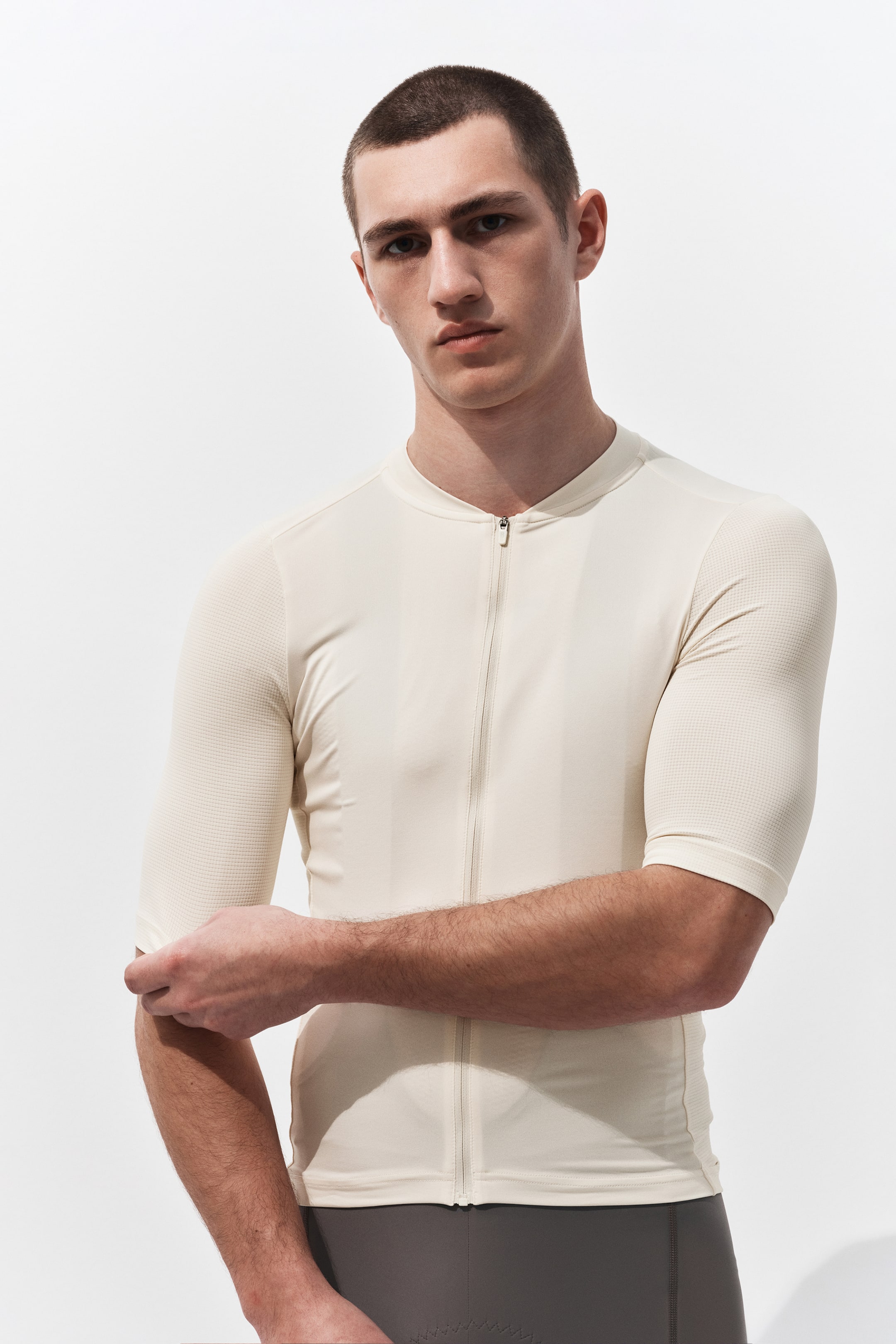 View larger image: A light cream cycling jersey is showcased, fitting snugly on a model facing forward. It features a low stand-up collar, a full-length front zipper, and short sleeves with a mesh-like textured fabric, contrasting with the smooth body.