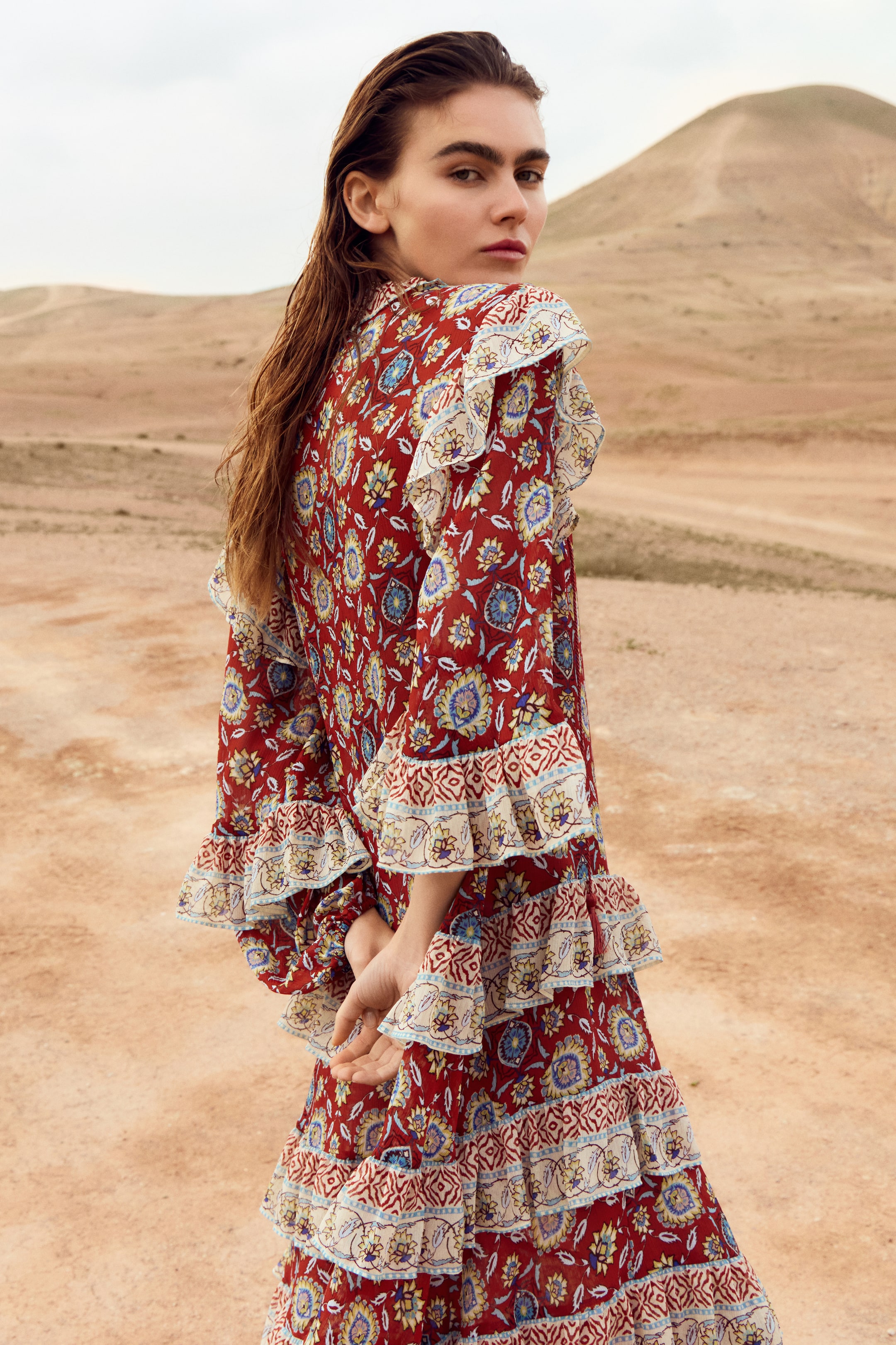 View larger image: The model is donning a long, A-line dress crafted in a red floral and geometric crinkled weave. She stands with her hands clasped behind her back. This dress offers a relaxed fit and showcases a round neckline, dropped shoulders, and long balloon sleeves with elasticated cuffs. The design is accentuated by wide flounces around the neckline, over the shoulders, on the sleeves, and in tiered flounces cascading down the skirt.