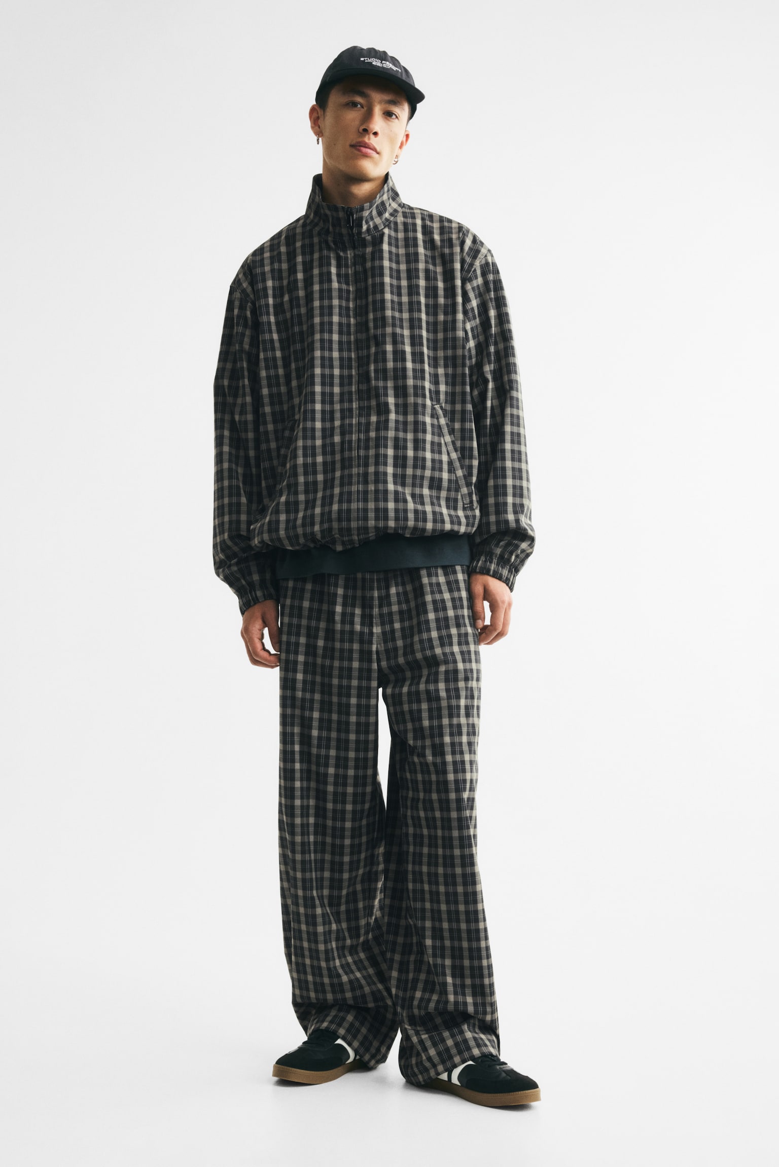 The trousers are shown from the front, showcasing a black and light brown check pattern. They boast an elasticated waistband and adjustable hems, complete with visible drawstrings and cord locks at the ankles. - 1