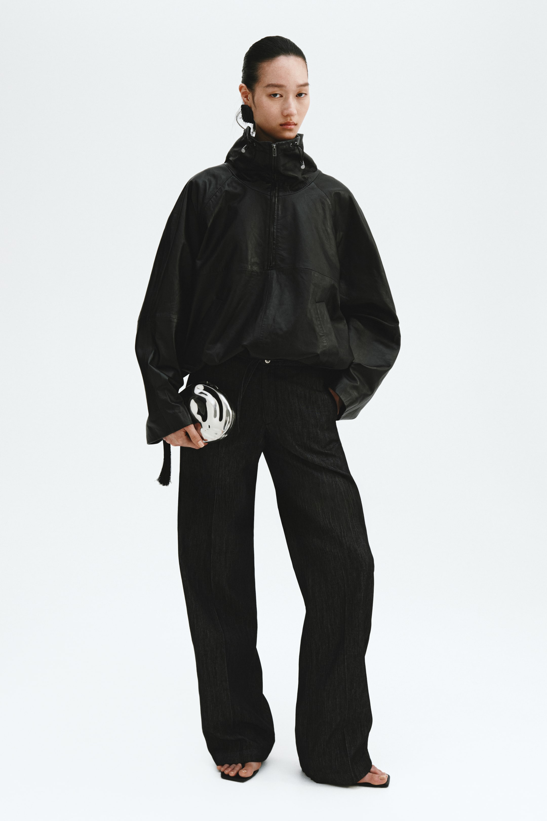 View larger image: A black textured anorak jacket featuring a high zippered collar and drawstring hem is paired with dark, wide-leg textured pants. The ensemble is accessorized with a silver, abstract-shaped clutch adorned with a black tassel, complemented by black square-toe open-toe sandals and a black sculptural earring.