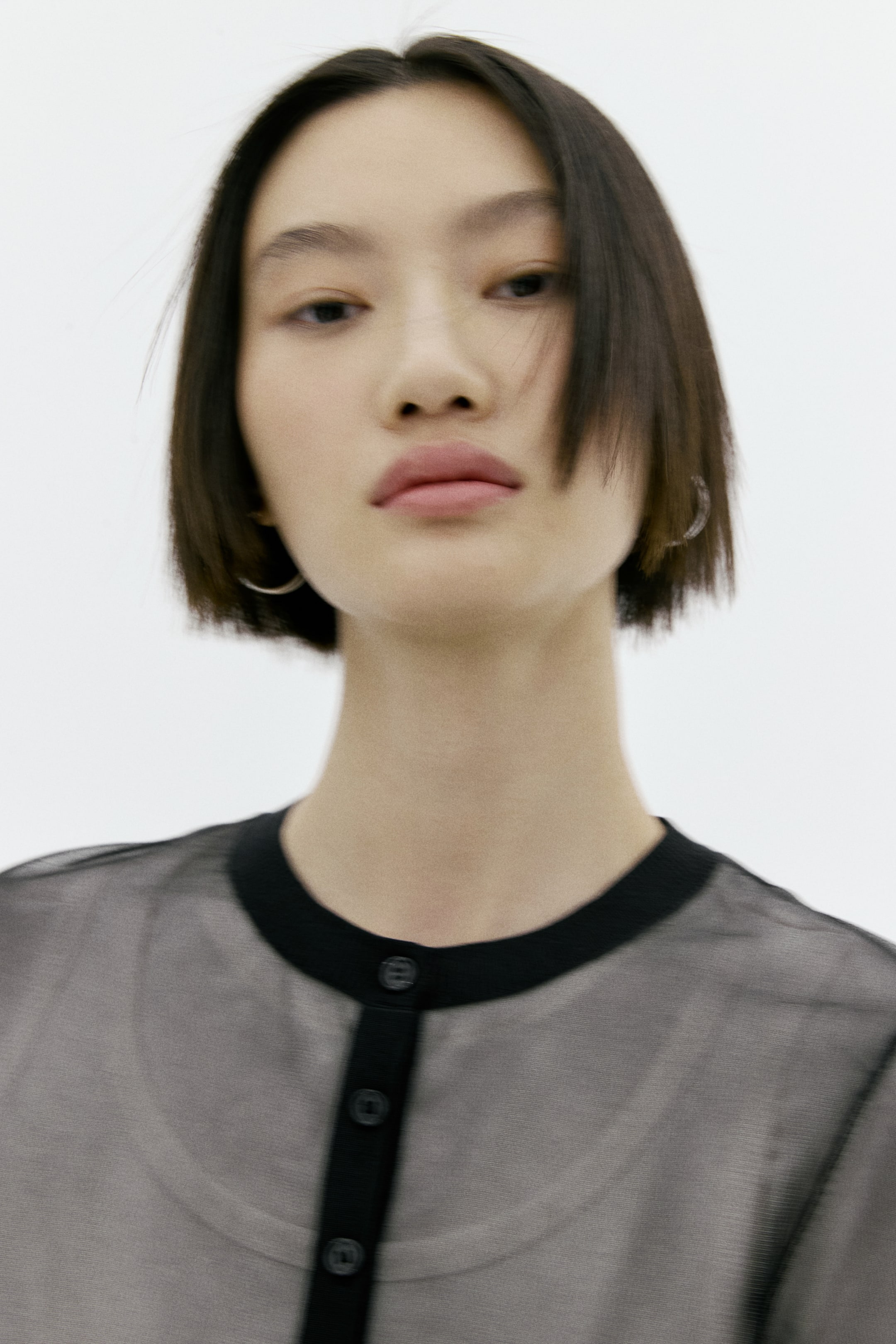 이미지 확대 보기: 시스루 블랙 카디건은 라운드 넥과 버튼 여밈 플래킷을 갖추고 있으며, 두 부분 모두 솔리드 블랙 원단으로 트리밍되어 있습니다. 여러 개의 블랙 버튼이 앞면을 고정하고 있으며, 밝은 톤의 이너셔츠가 시스루 소재 아래로 보이면서 제품의 투명함을 강조합니다.