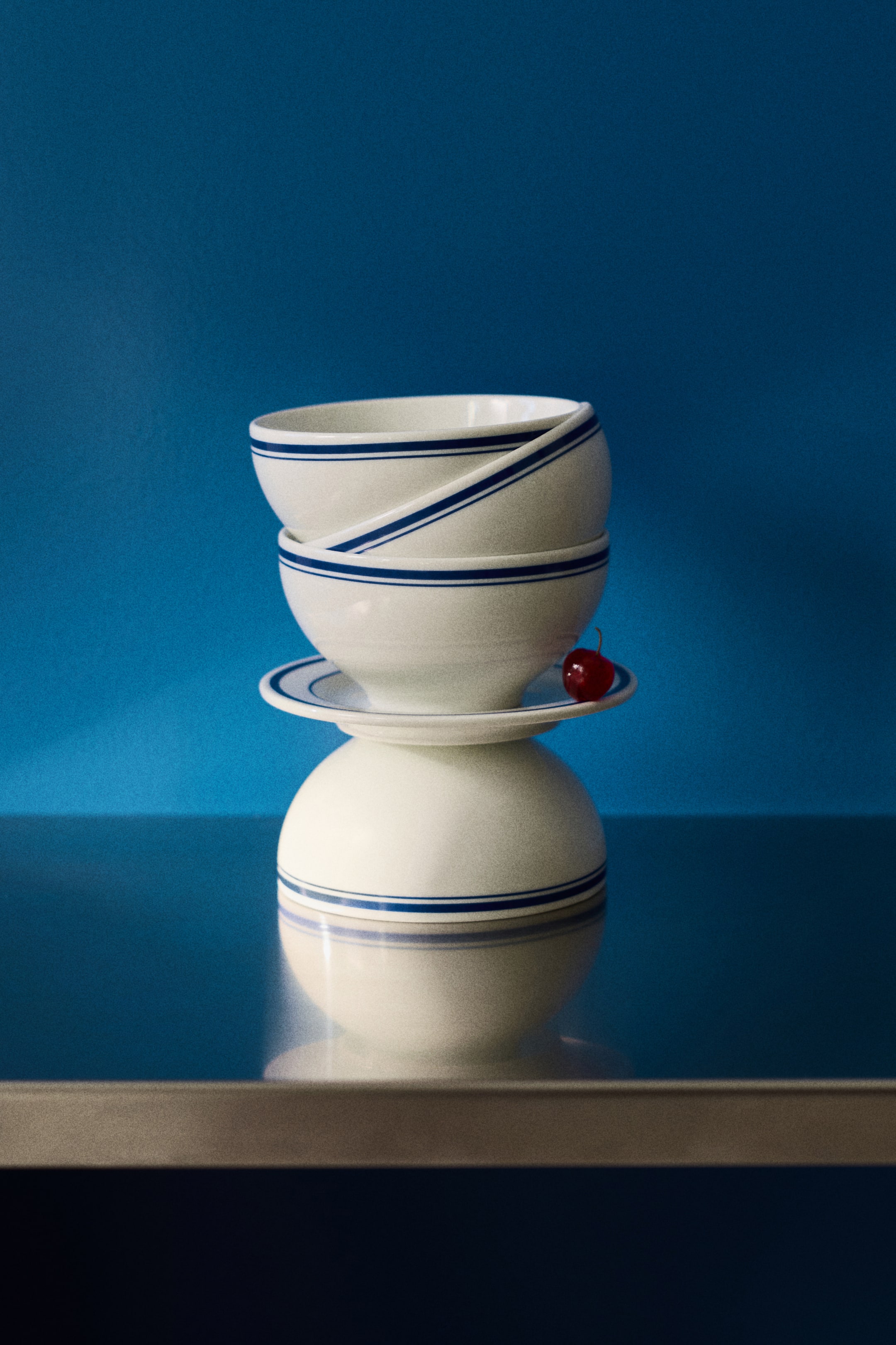 View larger image: Two white porcelain bowls, each adorned with double blue stripes near the rim, are stacked on a matching saucer. The saucer is placed on an inverted bowl of the same design, with a red cherry sitting on top.