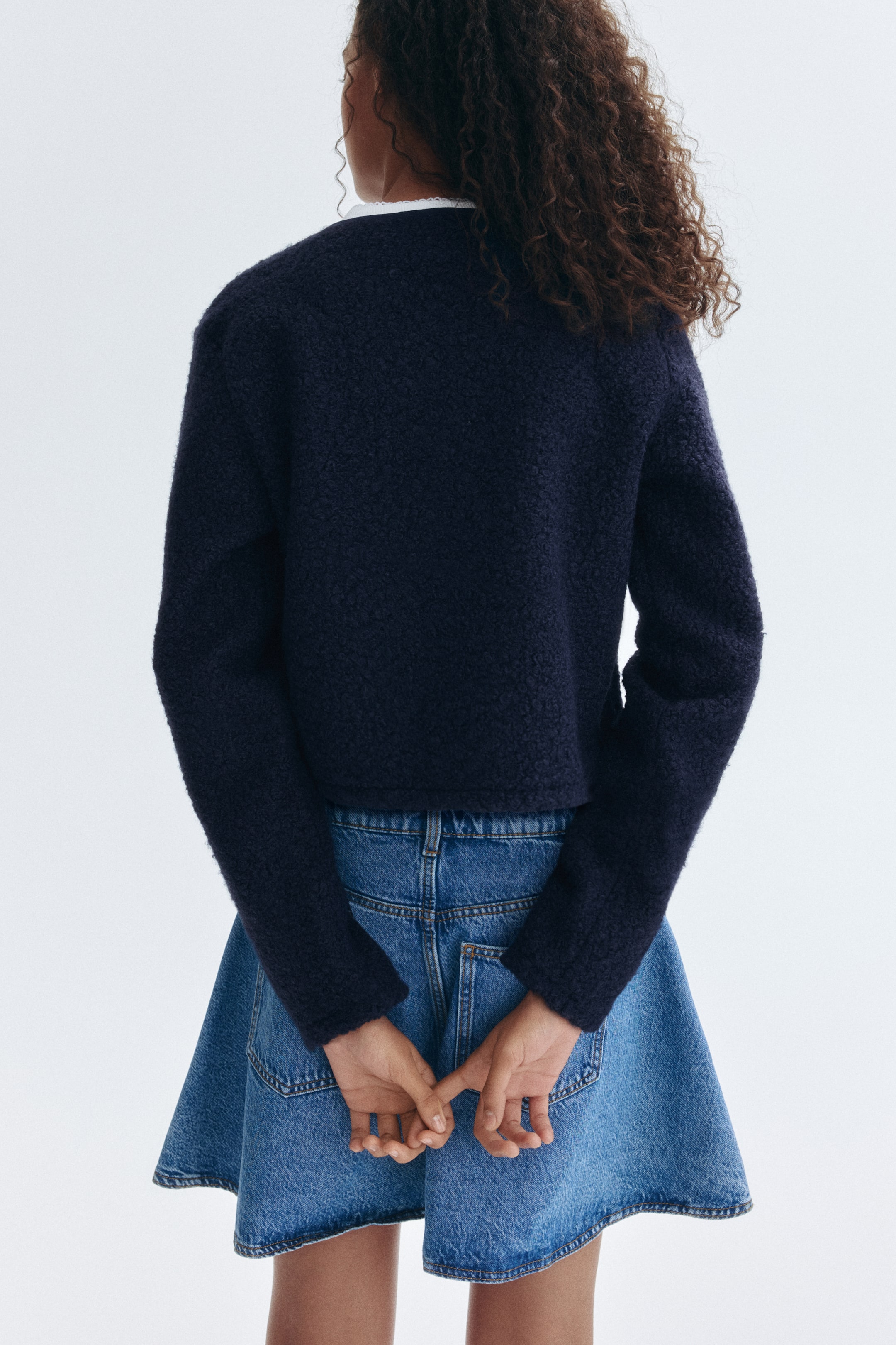 View larger image: A dark blue, textured knit cropped jacket is being worn by the model, with a white collar visible underneath. The model's hands are clasped behind her back, resting against a blue denim A-line skirt.