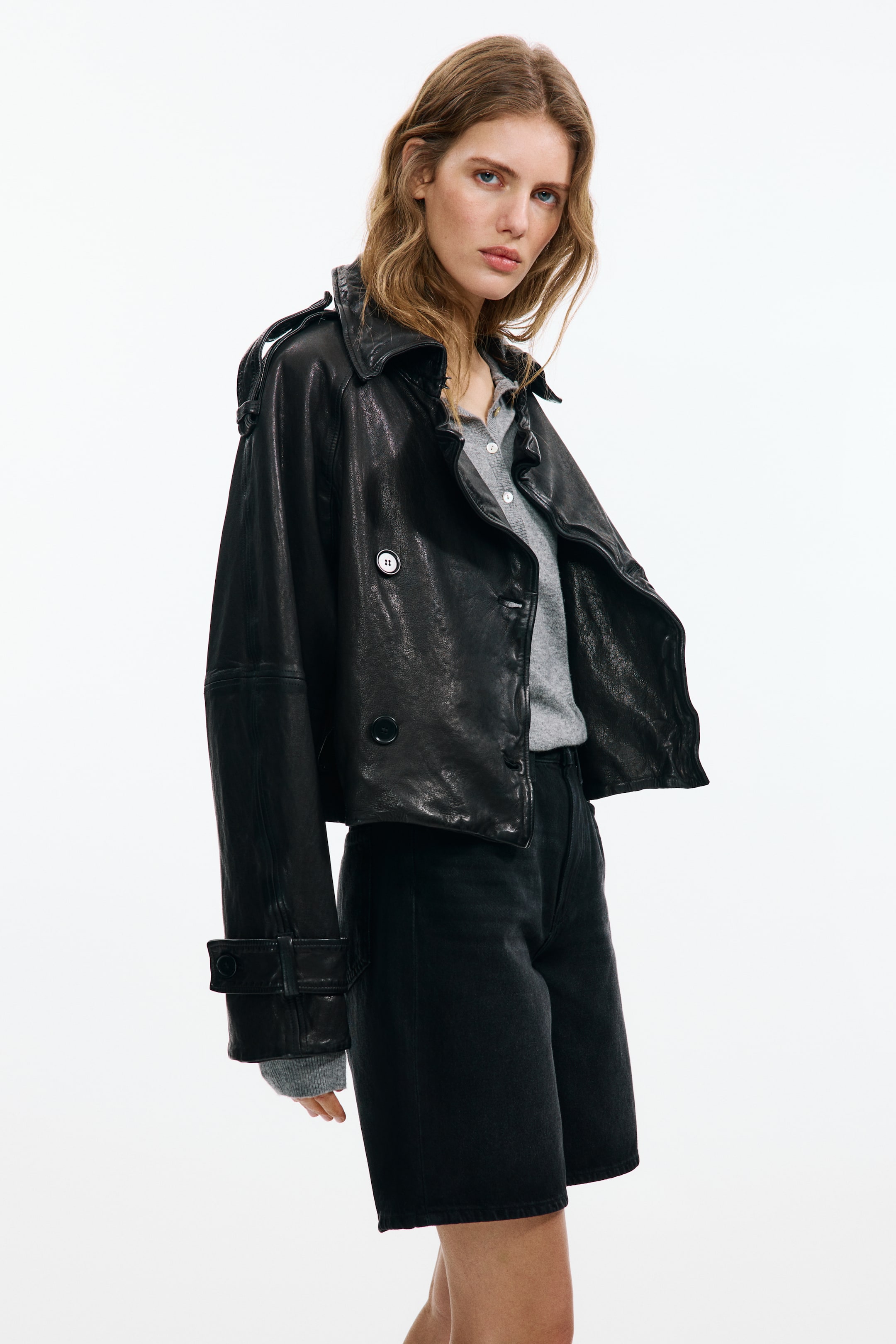 View larger image: A black leather cropped jacket is displayed from a slight front-left angle, highlighting a distressed texture, a double-breasted front with two visible buttons, and classic lapels. It also features shoulder straps and adjustable strap and button detailing on the cuffs, with a grey top visible underneath.