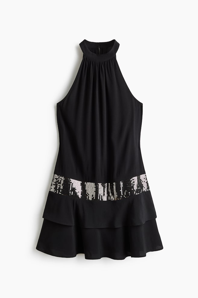 Una vista frontal de un vestido negro de cuello halter muestra un cuello alto fruncido y un diseÃ±o sin mangas. Una amplia banda horizontal de lentejuelas plateadas brillantes rodea la parte inferior de la cintura y da paso a un bajo con volantes de dos niveles. - 1