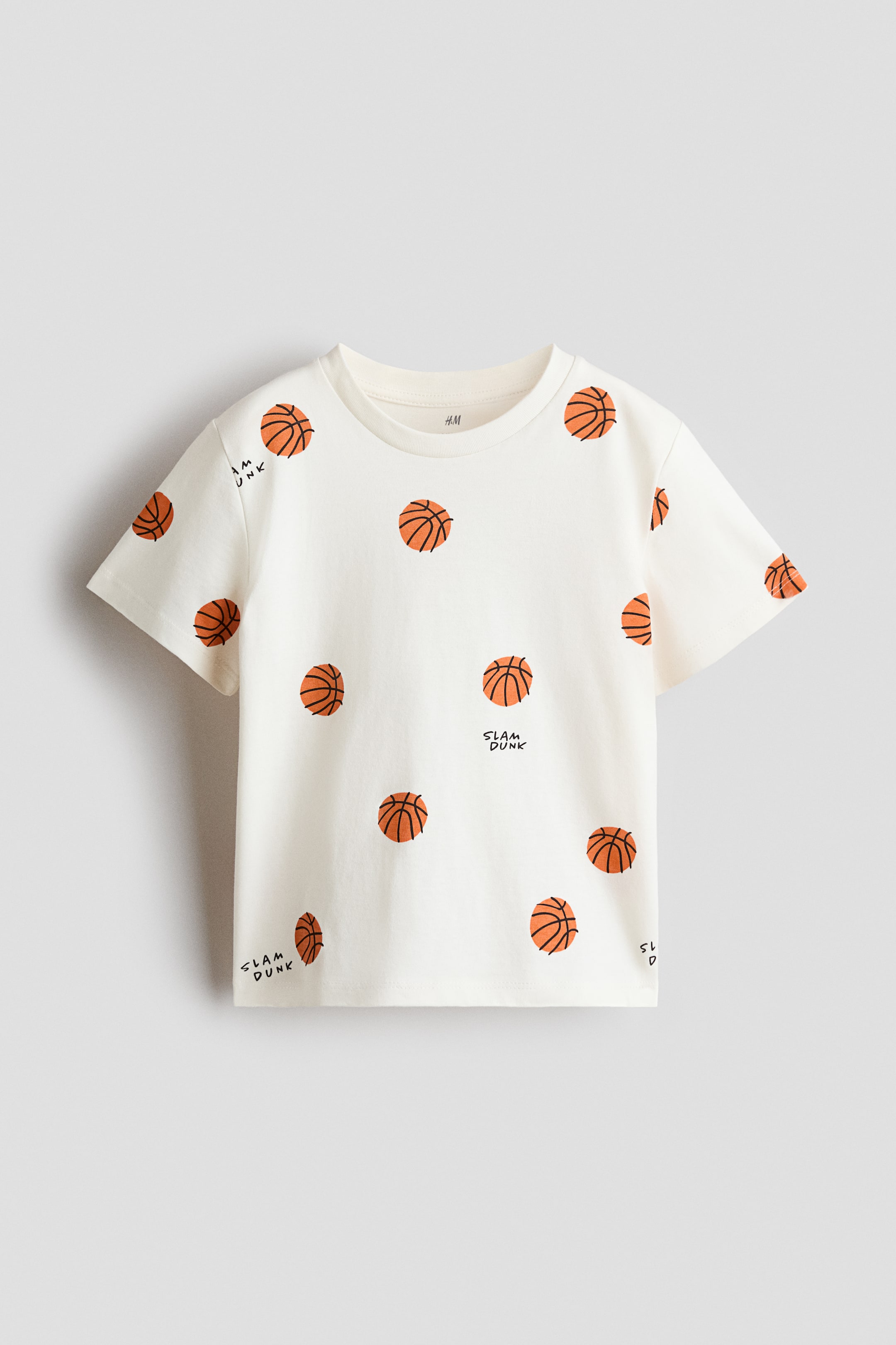 拡大画像を表示: 柔らかなコットンジャージー素材のTシャツが正面から紹介されています。オフホワイトのカラーに、オレンジ色のバスケットボールと「SLAM DUNK」テキストが全面にプリントされています。リブ付きのクルーネック、ドロップショルダー、ストレートカットの裾が特徴です。