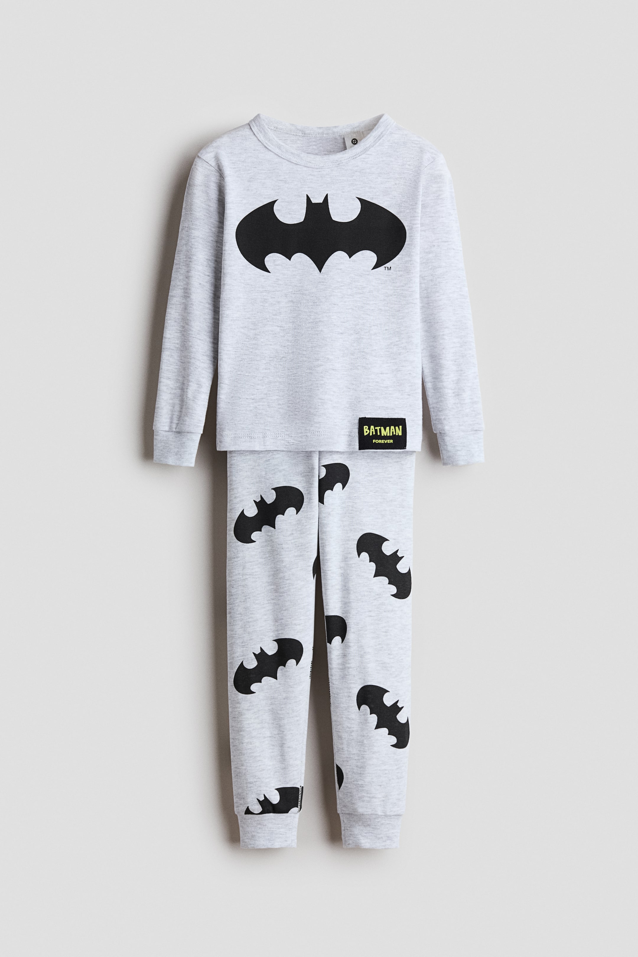View larger image: A light gray marl cotton jersey pajama set features a long-sleeved top and matching bottoms. The top displays a large black Batman logo on the chest, a small black and yellow 'Batman Forever' label near the hem, a round neckline with a narrow trim, and ribbed cuffs. The bottoms showcase an all-over black Batman logo print and ribbed hems.