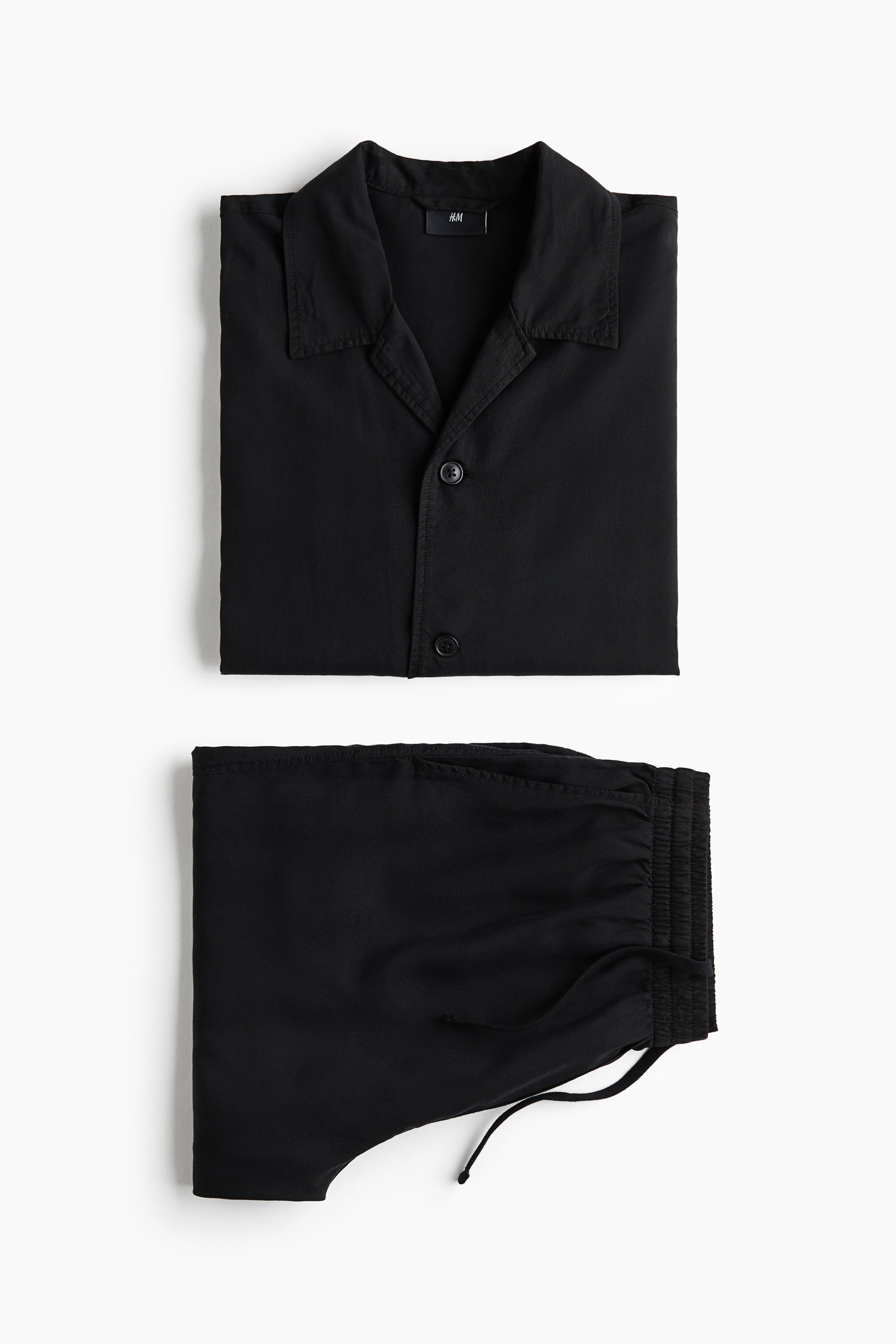 View larger image: A black resort shirt and matching shorts set is laid flat, displayed from the front. The short-sleeved shirt features a resort collar, a French front with two visible buttons, and a straight-cut hem, while the shorts have an elastic waist with visible drawstrings.