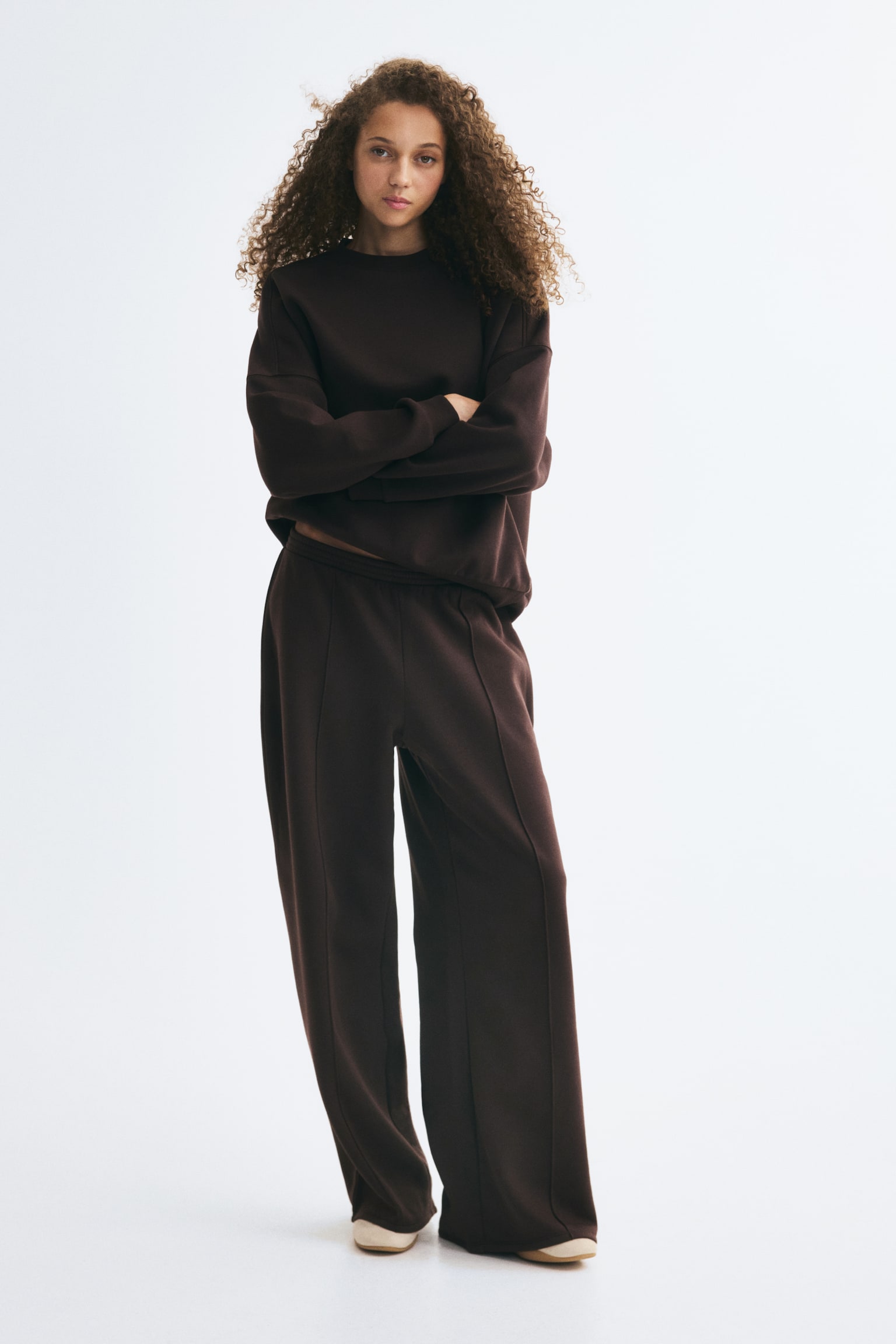 A dark brown two-piece loungewear set is hanging, facing the front, featuring an oversized crew-neck sweatshirt with dropped shoulders, ribbed cuffs, and a ribbed hem. The matching wide-leg trousers are hanging underneath, showing vertical centre seams running down each leg. - 1