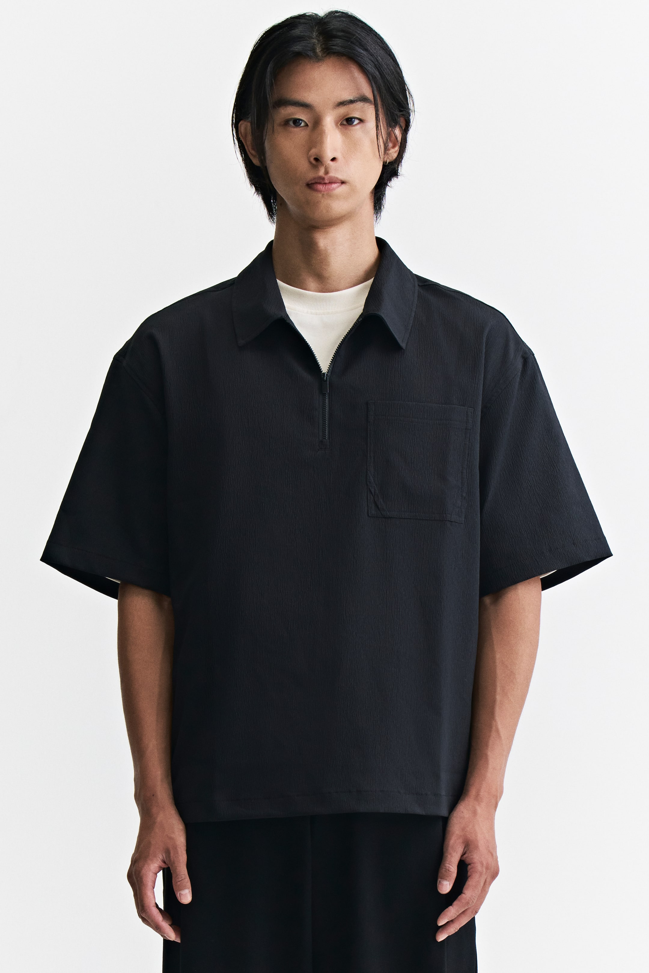 View larger image: A black polo shirt in a crinkled weave is worn by the model, featuring a relaxed fit, a collar with a half-zip closure, short sleeves, and a single chest pocket. The shirt is layered over a white crew-neck t-shirt.
