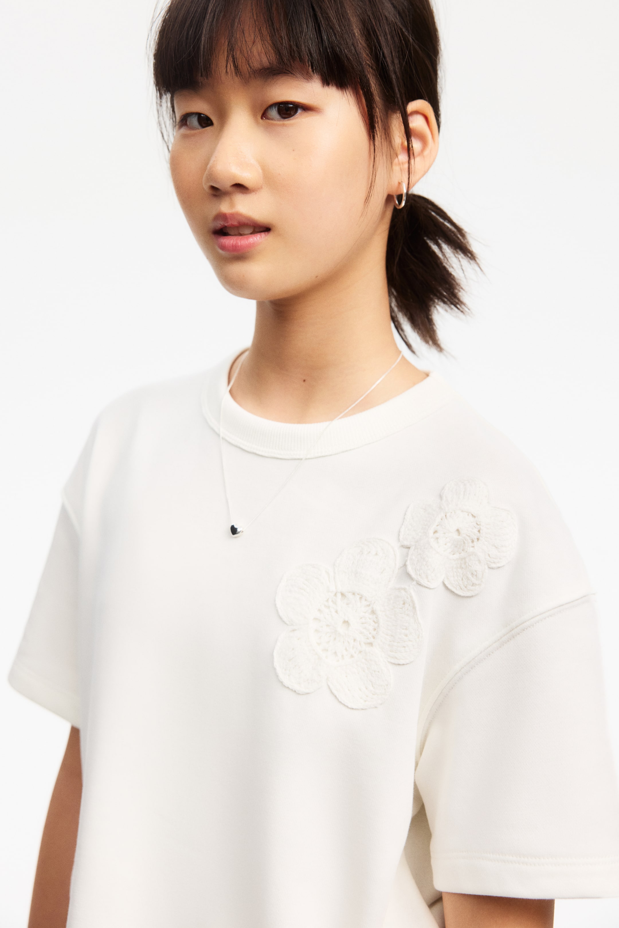 Agrandir l'image: Un top blanc à manches courtes présente un col rond et deux appliqués floraux en crochet blancs sur le côté gauche de la poitrine. Un fin collier en argent avec un petit pendentif en forme de cœur noir repose sur l'encolure du top.