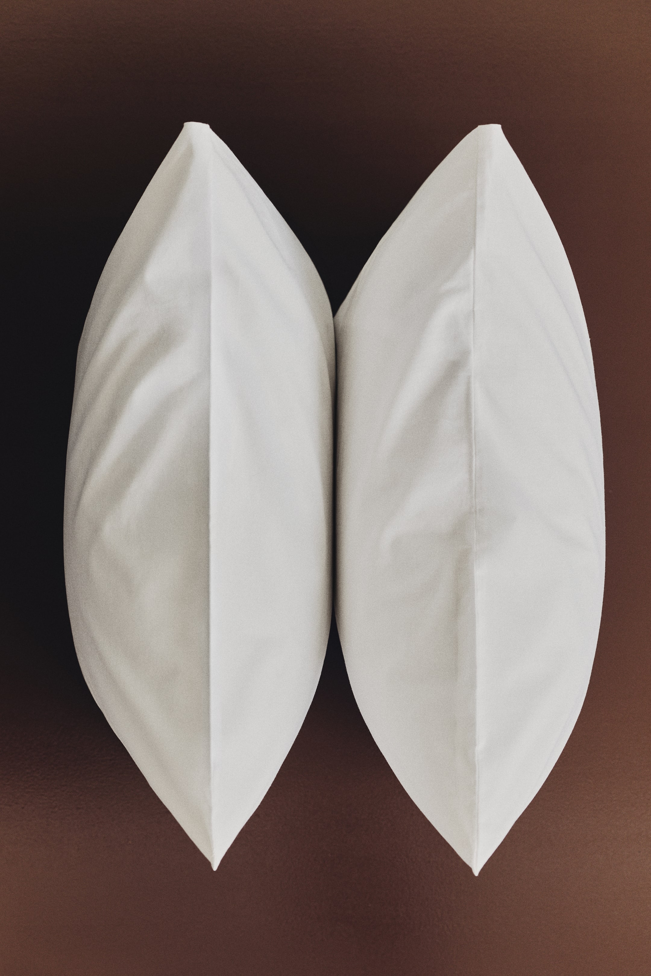 Ampliar la imagen: Un par de fundas de almohada blancas en un tejido de algodón suave se encuentran de pie, una al lado de la otra, con aperturas tipo sobre visibles como tela doblada en los extremos superior e inferior.