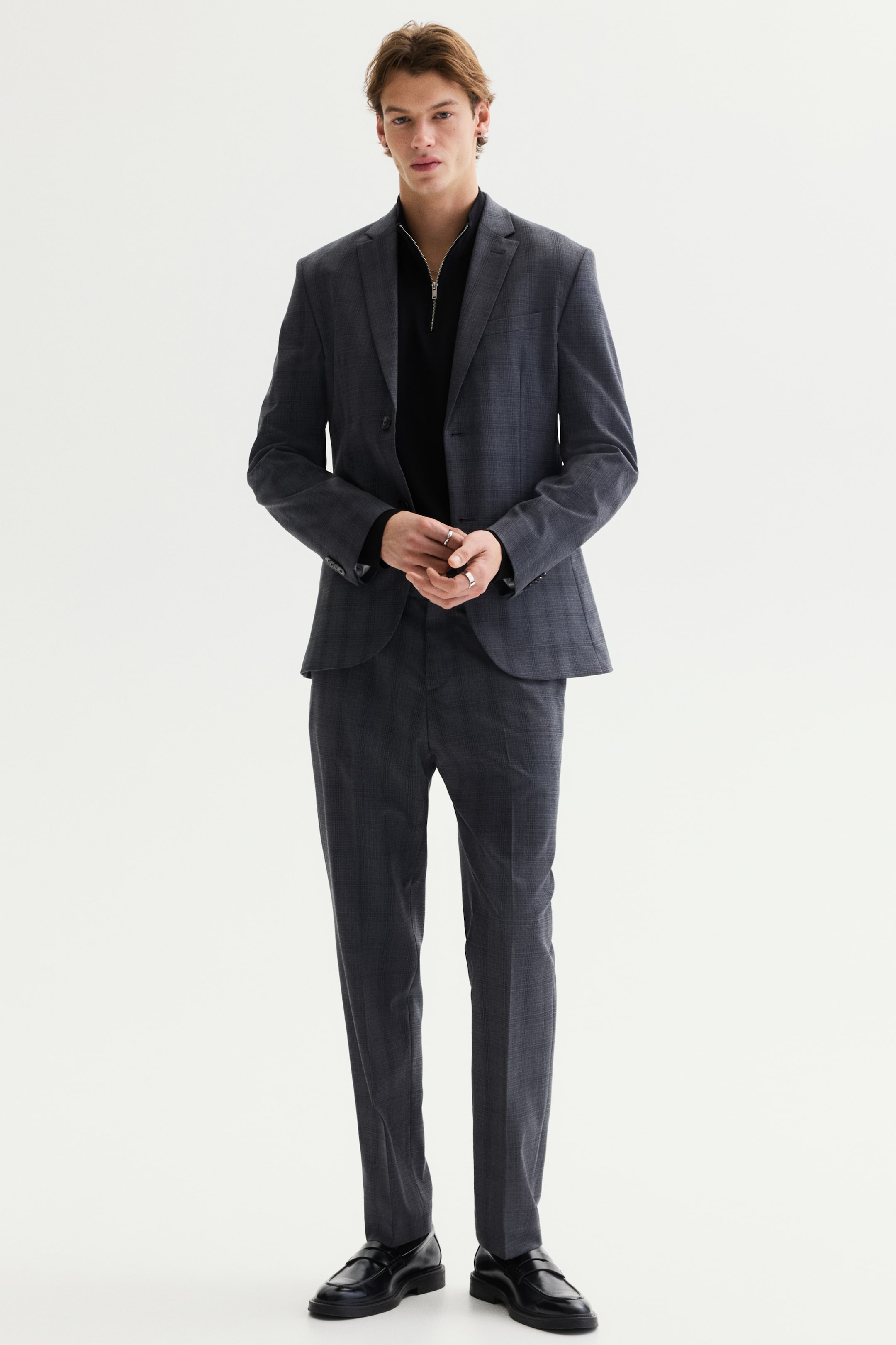 View larger image: A dark grey patterned suit is showcased from a frontal perspective. The single-breasted jacket, featuring notch lapels, a two-button closure, and flap pockets, is layered over a black quarter-zip top, complementing matching slim-fit trousers worn with black loafers.