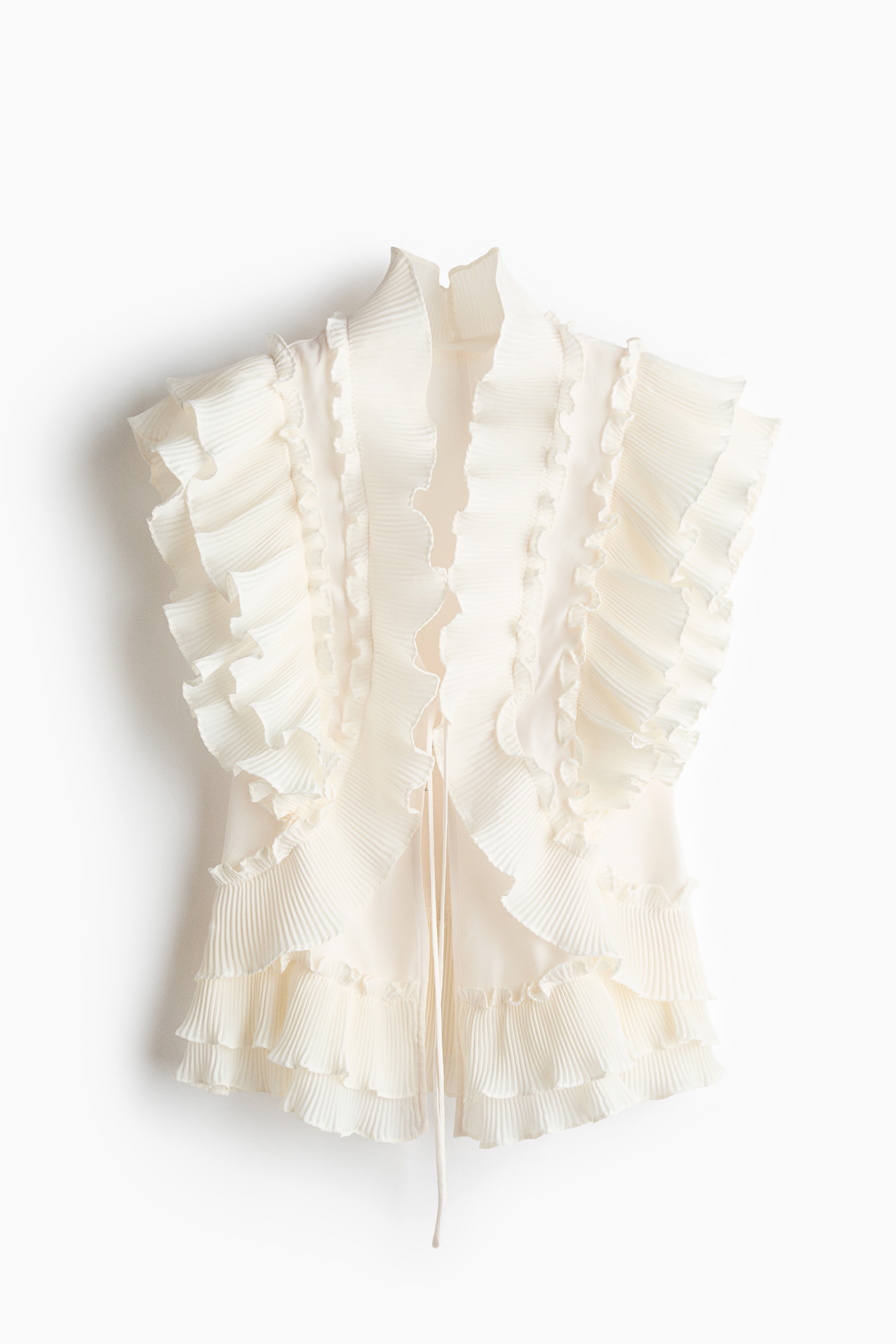 A cream-colored sleeveless blouse featuring tiered flounces and a frill collar is being worn by the model over a black bra. The open-front blouse is secured with narrow ties and paired with black trousers, while the model is holding a small metallic chain handbag.