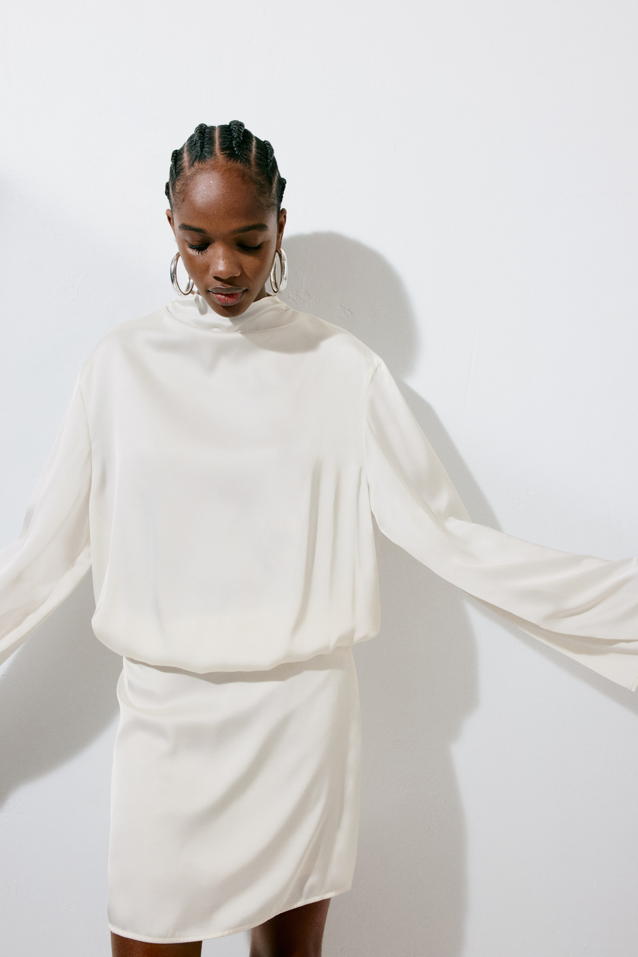 View larger image: The model is donning a short, off-white satin dress that boasts a stand collar, a relaxed bodice gracefully draping at the waist, and dropped shoulders complemented by long, wide sleeves. With arms extended outward, the model highlights the sleeves and the fitted skirt that elegantly falls above the knee.