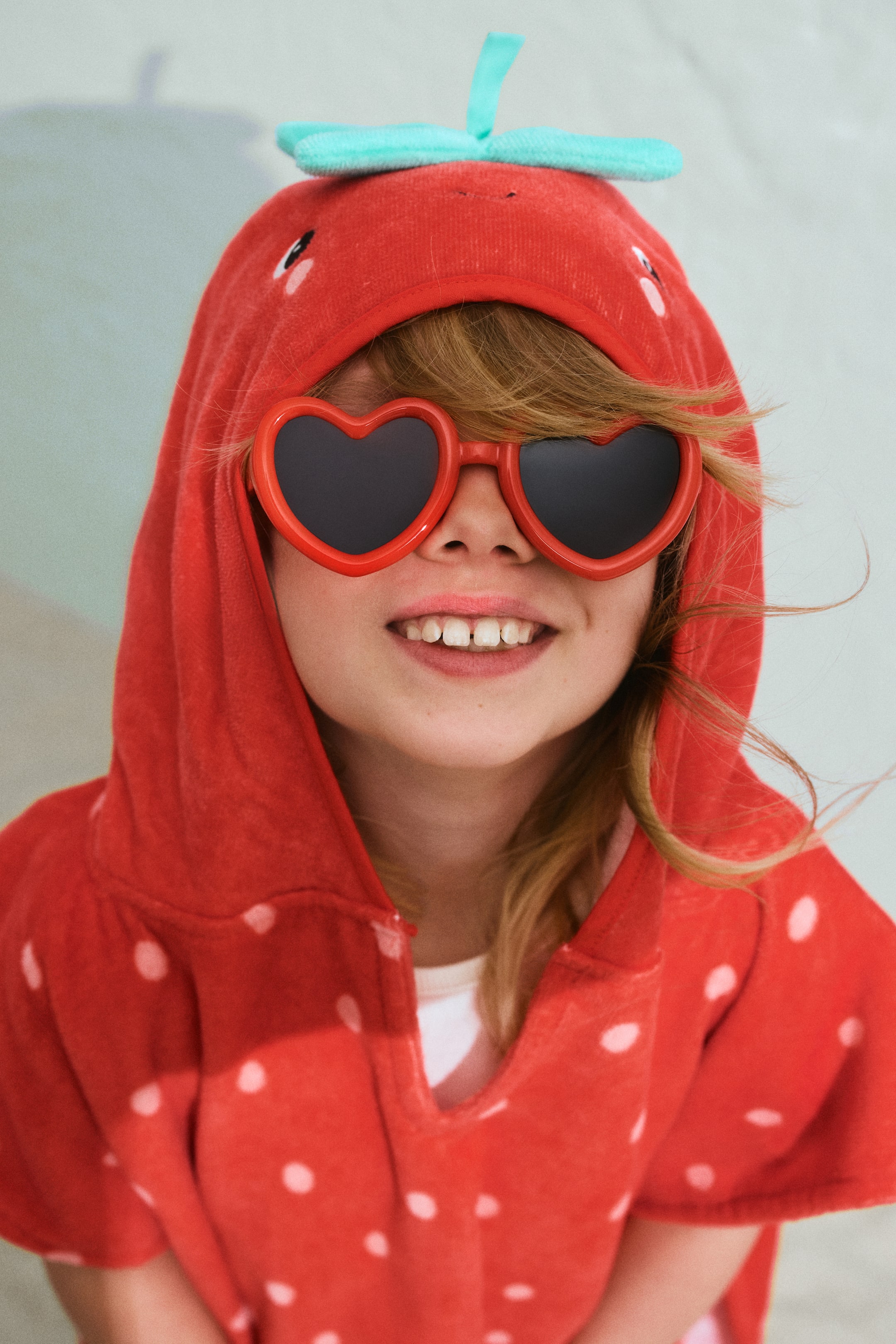 Grotere afbeelding bekijken: Een rood badstof capuchonponcho wordt gepresenteerd vanuit een vooraanzicht, met witte stippen en een aardbeivormige capuchon met een lichtblauwe steel en zwart geborduurde zaadjes. Rode hartvormige zonnebrillen worden gedragen, omlijst door de capuchon.