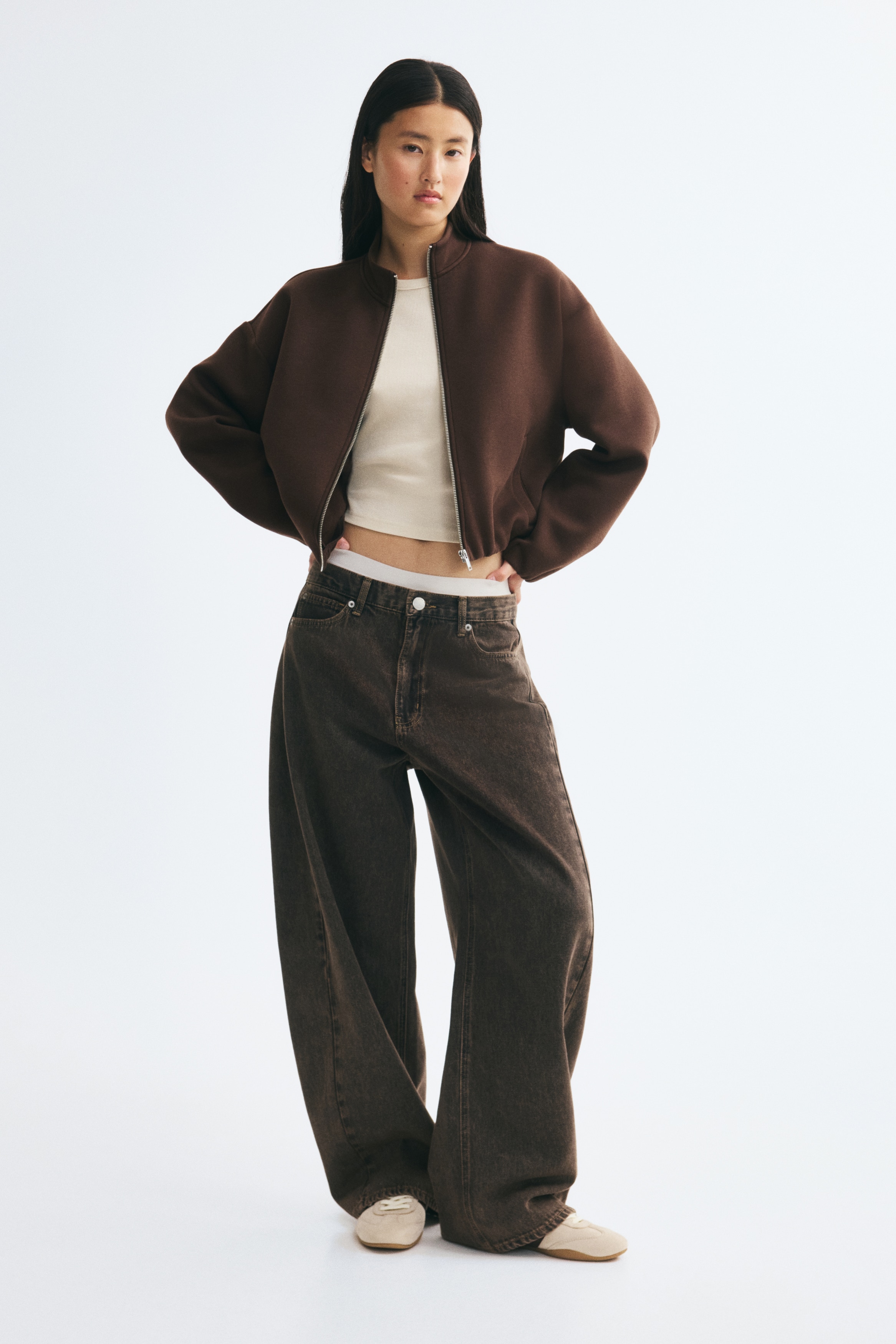 A dark brown zip-up sweatshirt jacket is shown from the front, featuring a high stand-up collar and a full-length silver zipper closure. It is designed with dropped shoulders, wide sleeves with gathered cuffs, vertical welt pockets, and an elasticated hem that creates a slightly cropped silhouette.