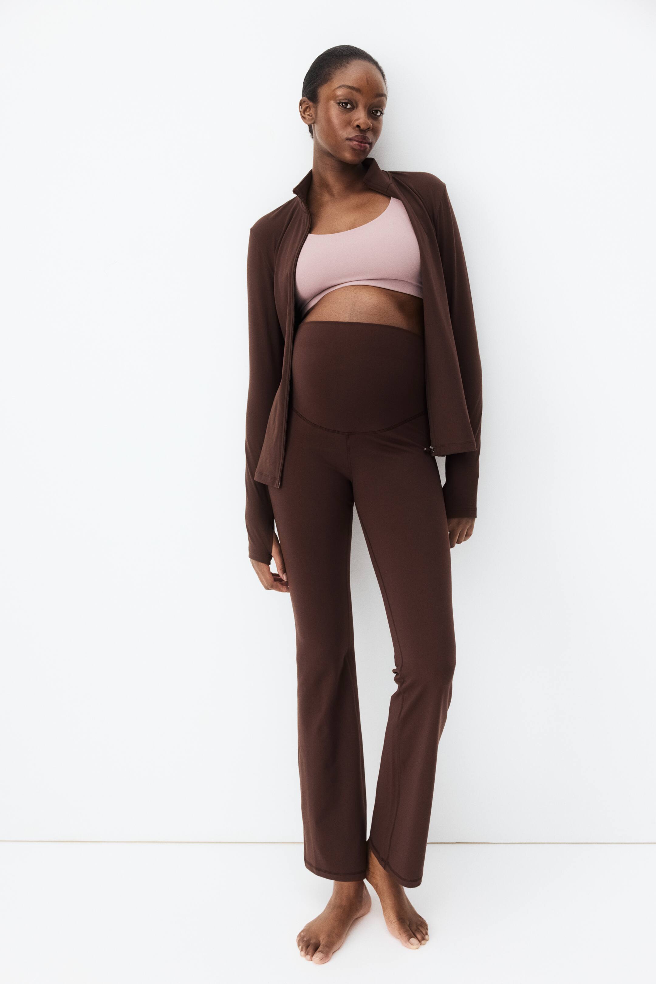 Ampliar la imagen: Una chaqueta marrón de manga larga está abierta, revelando un brasier deportivo rosa claro y unos pantalones maternales marrones de talle alto. Los pantalones presentan una cinturilla ancha y de soporte y una abertura amplia en la pierna.