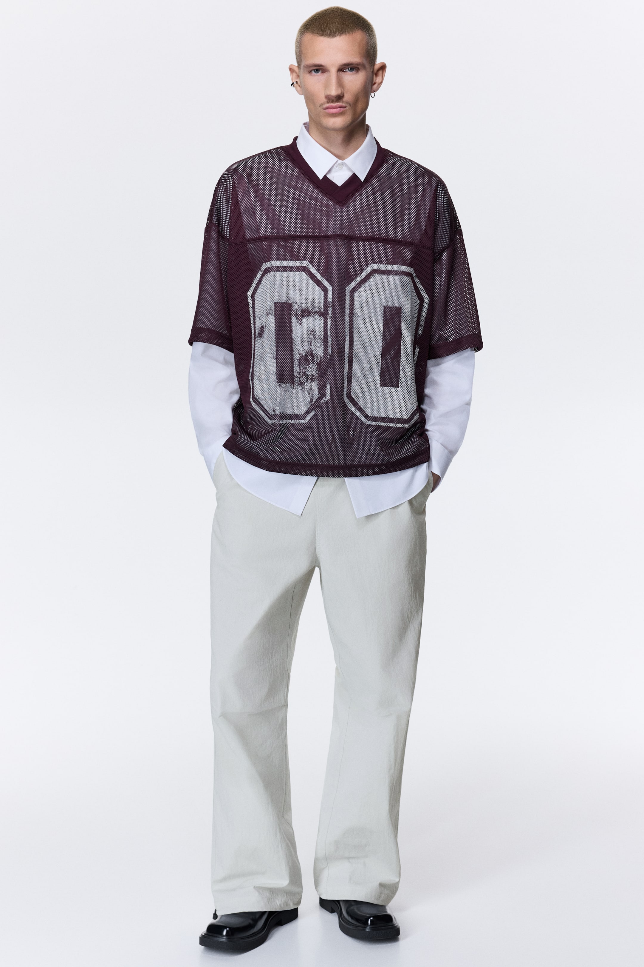 Xem ảnh lớn hơn: Một chiếc áo jersey lưới màu burgundy với cổ chữ V, viền cổ gân và tay ngắn, nổi bật với số “00” màu trắng cỡ lớn, hiệu ứng cũ xước ở mặt trước. Áo được phối layer bên ngoài một chiếc áo sơ mi cổ bẻ màu trắng.