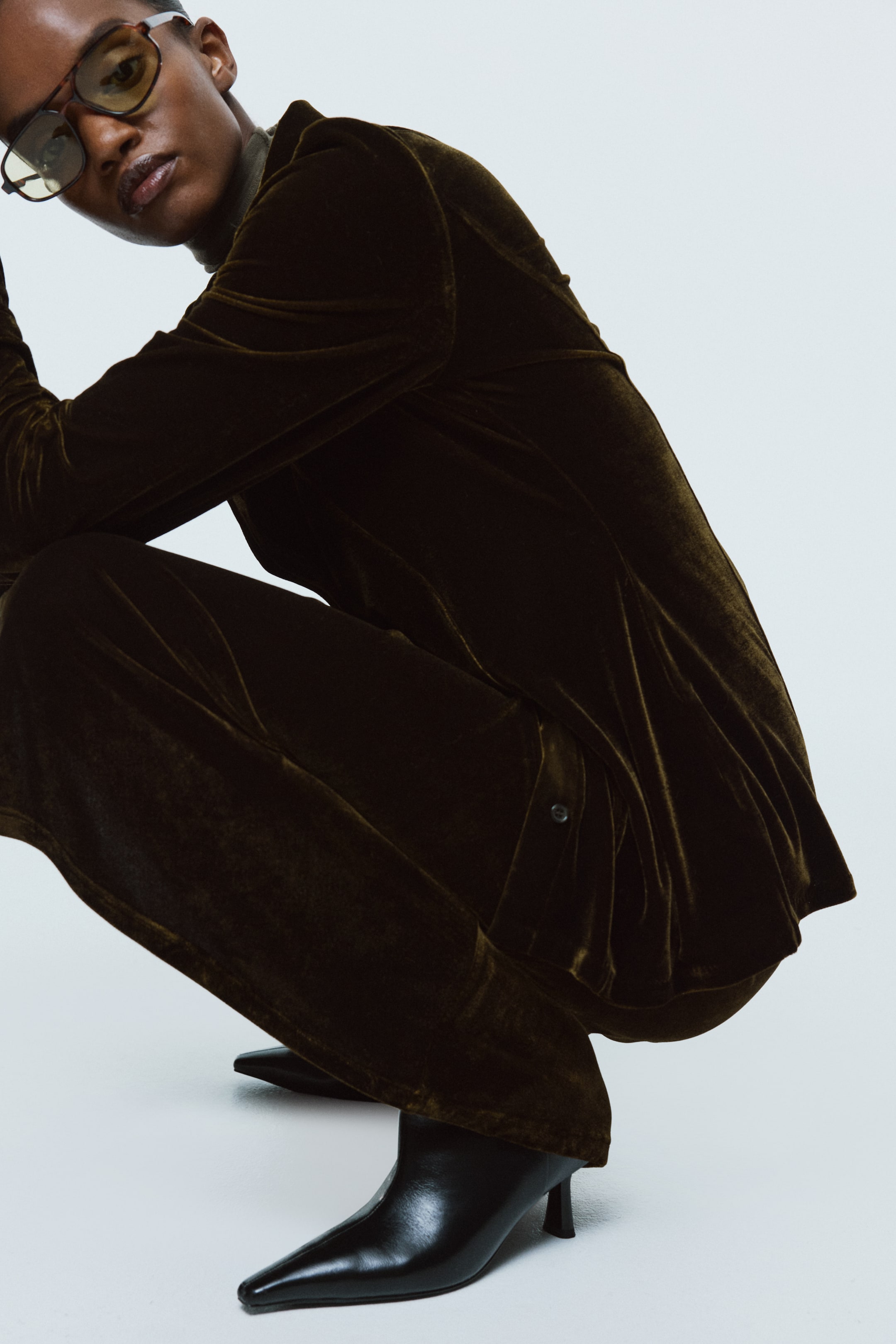 View larger image: A dark olive velvet long-sleeved top, featuring a visible side button, is being layered over a lighter turtleneck and paired with matching wide-leg trousers. Black pointed-toe ankle boots with kitten heels are seen below the trousers, with the velvet fabric softly draping and reflecting light as the wearer is crouching.