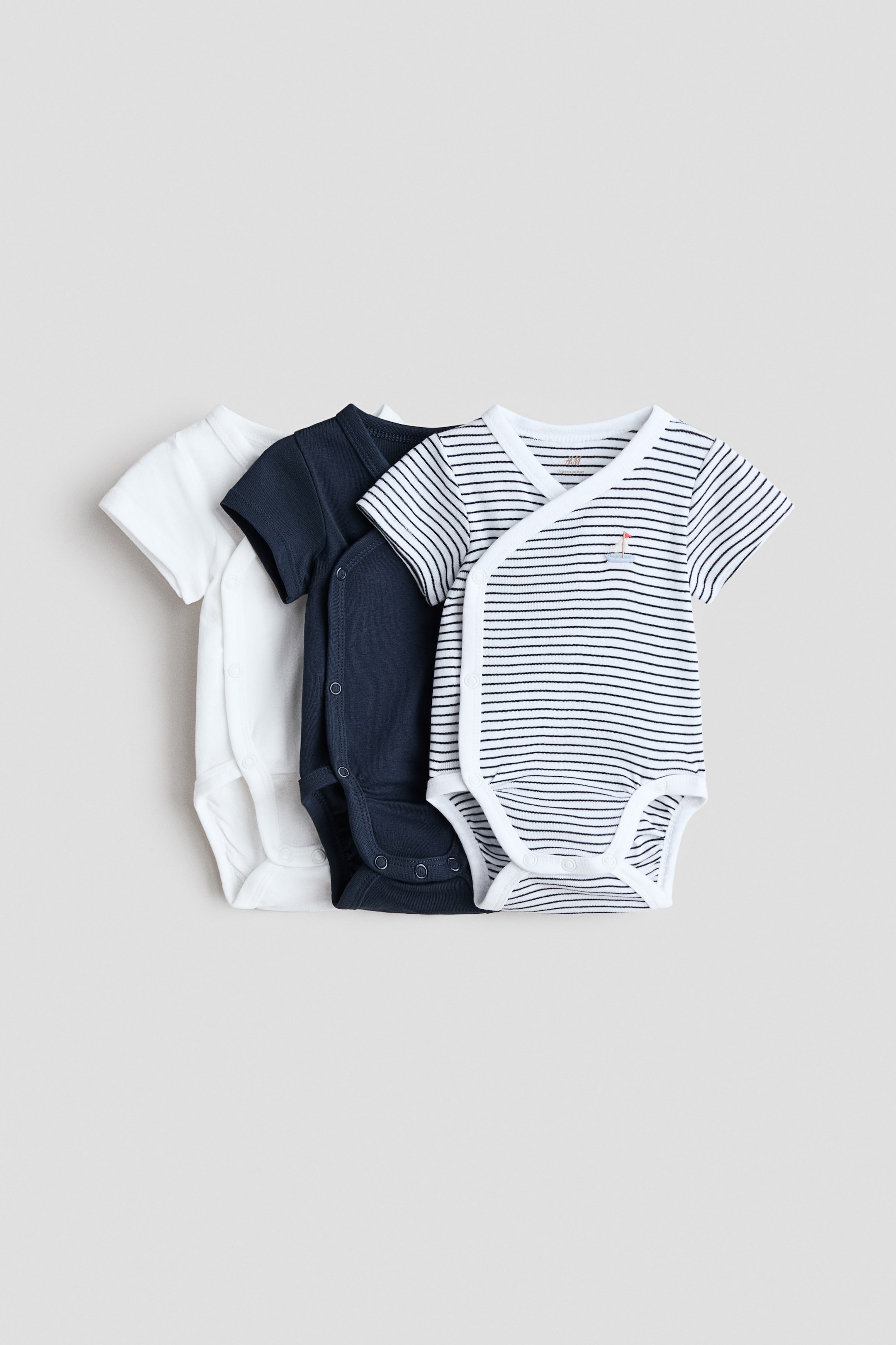 View larger image: Presenting a set of three short-sleeved wrapover bodysuits crafted from soft cotton jersey. The front bodysuit showcases elegant white and navy horizontal stripes, accented by a delicate sailboat embroidery. Behind it, a solid navy bodysuit and a solid white bodysuit layer seamlessly. Each piece is designed with convenient press-studs along one side and at the crotch for easy dressing.