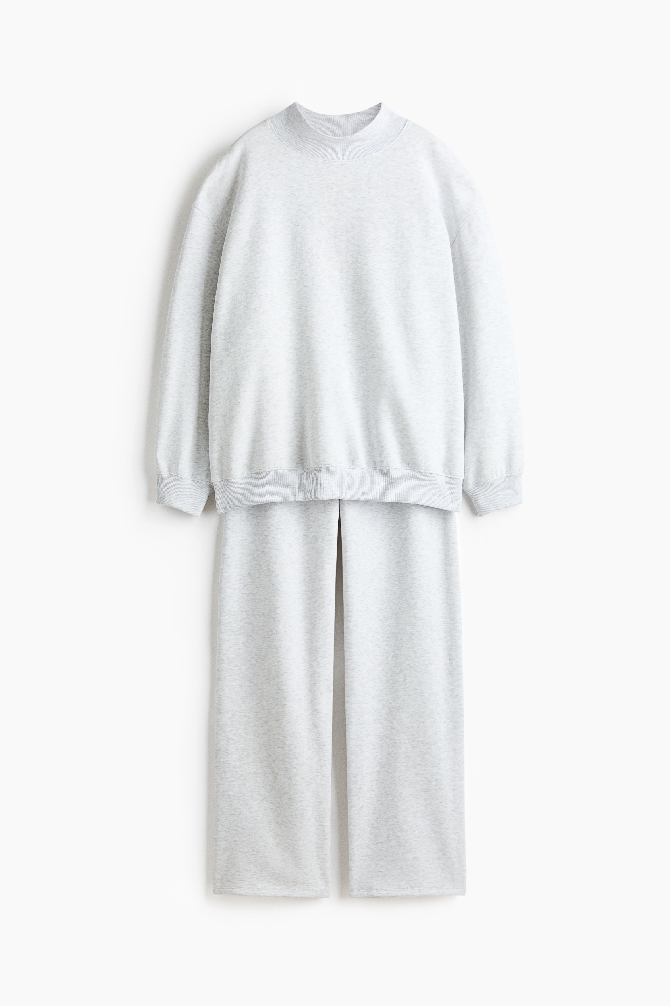 A matching light grey fleece sweater and track pants set is worn by the model, who is sitting cross-legged. The sweater features a high, ribbed neck, dropped shoulders, and ribbed cuffs, while the straight-leg track pants are styled with white and beige sneakers.