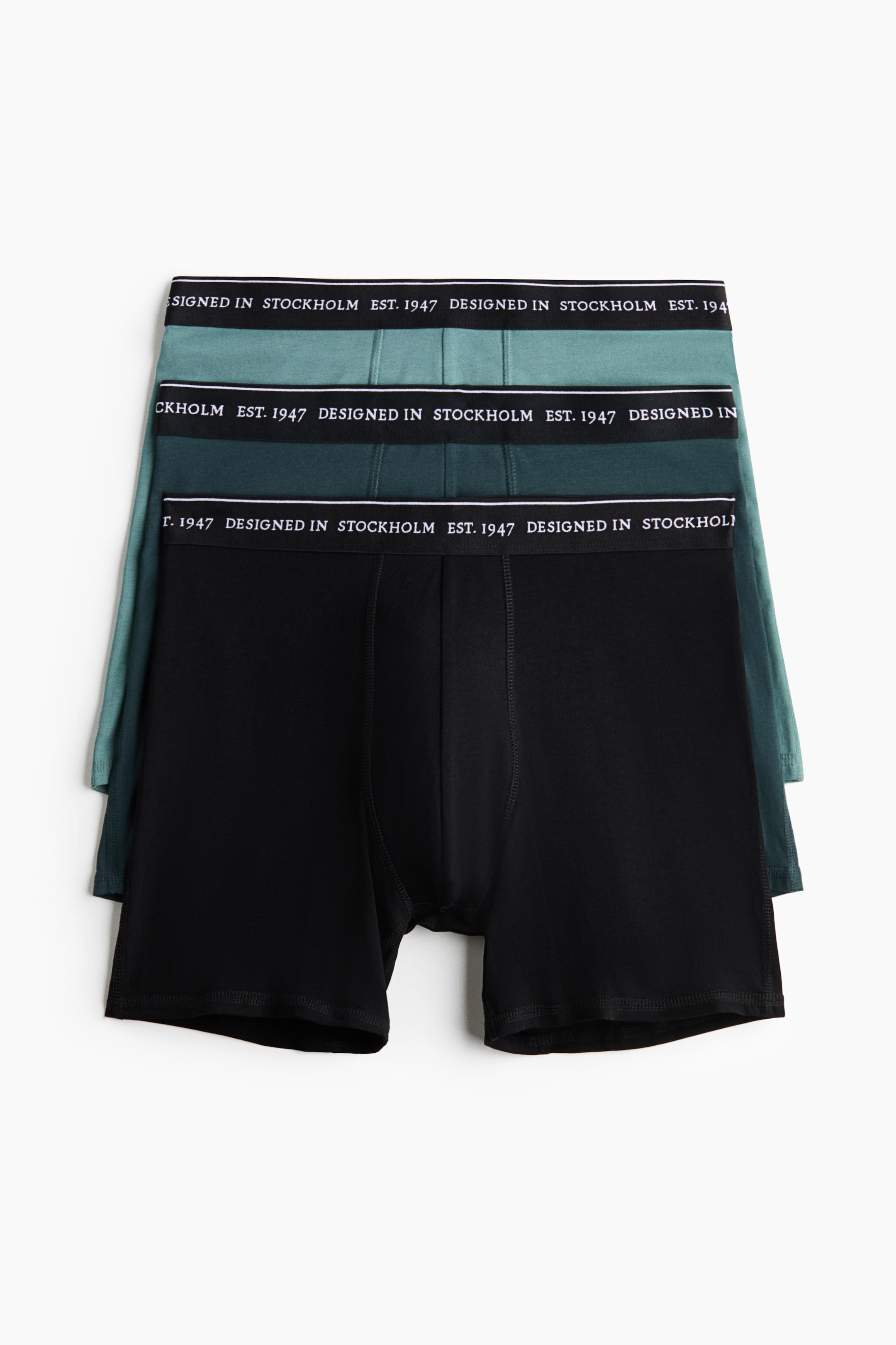 View larger image: Three pairs of men’s boxer briefs are laid flat, stacked with the front facing forward. The top pair is black, the middle pair is dark teal, and the bottom pair is light teal. Each pair features a black elastic waistband with white text reading "DESIGNED IN STOCKHOLM EST. 1947".