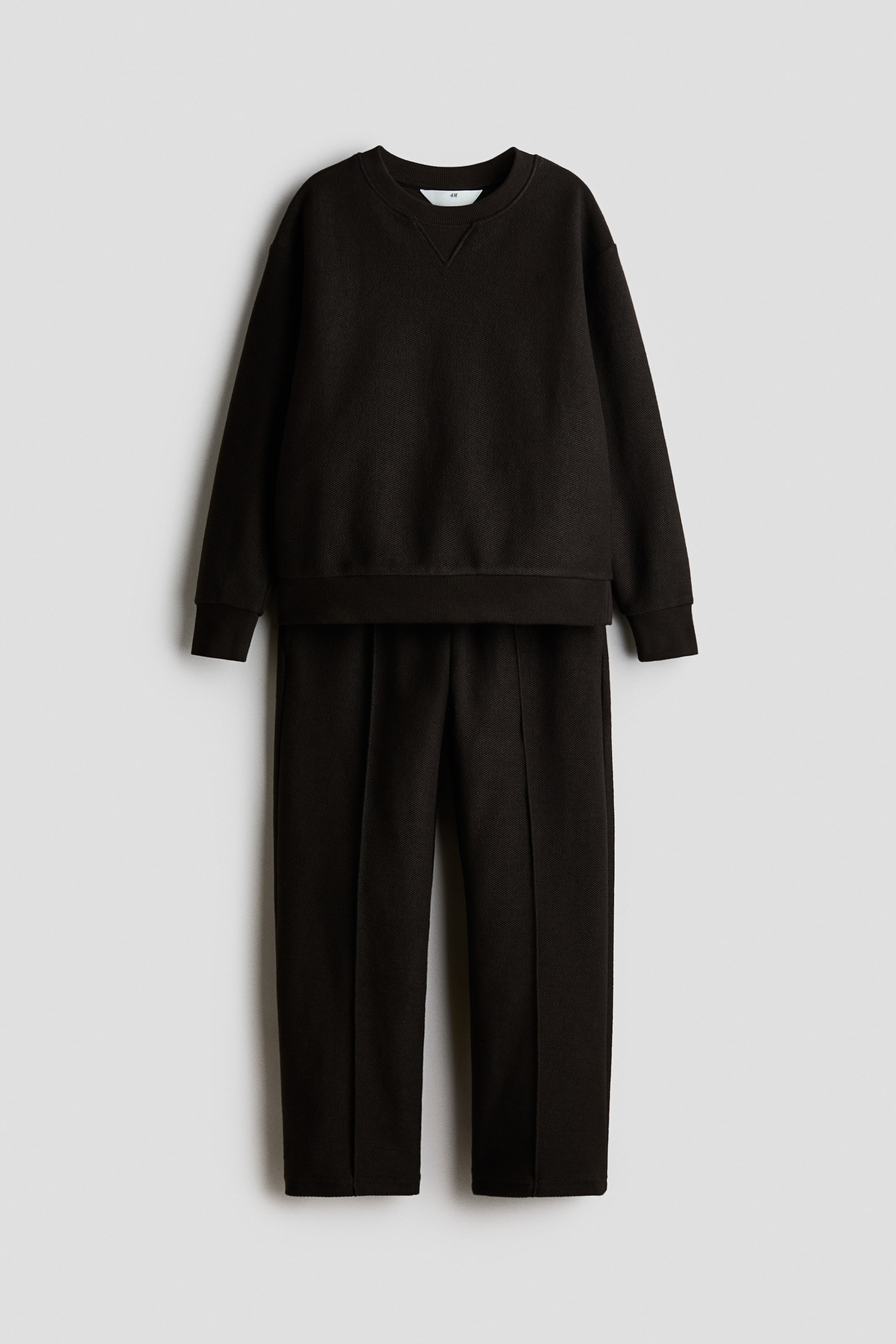 View larger image: A dark brown sweater and track pants set in textured sweat fabric is shown from a clean front view. The sweater features a rib-trimmed crew neckline with a V-stitch detail, dropped shoulders, and ribbed cuffs, while the matching pants have straight legs with sewn-in creases running down the front.