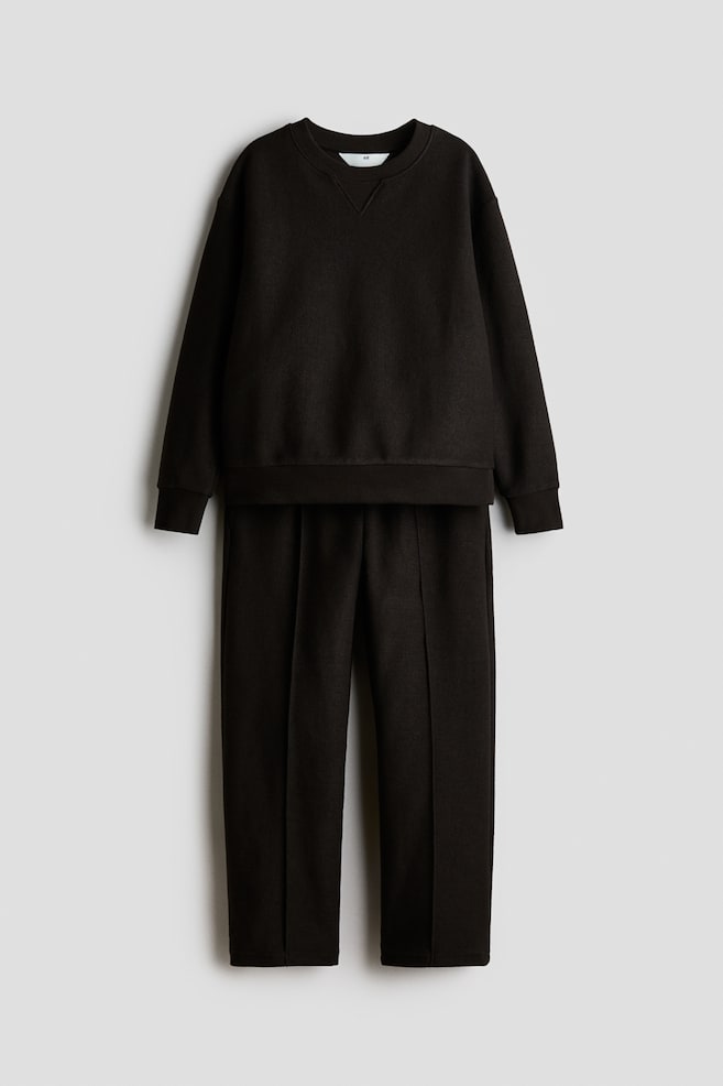 A dark brown sweater and track pants set in textured sweat fabric is shown from the front, with a clean, minimal look. The sweater features a round, rib-trimmed neckline with V-stitch detailing, dropped shoulders, and ribbed cuffs, while the matching pants feature straight legs with sewn-in front creases. - 1