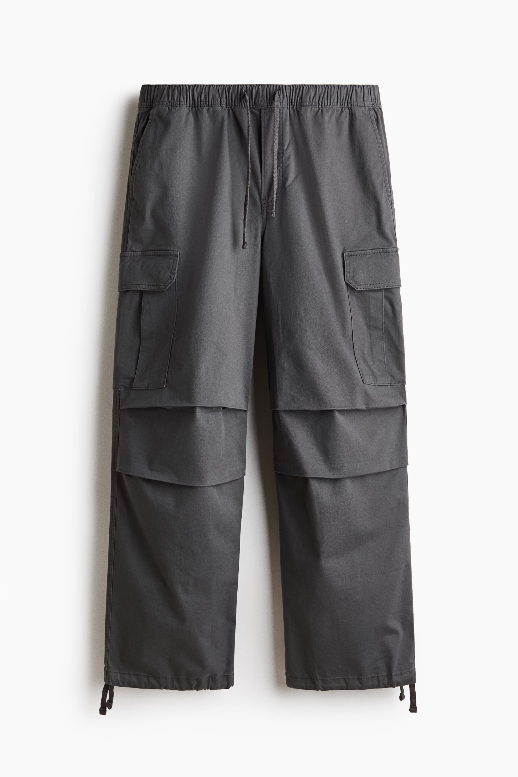 View larger image: Dark grey cargo pants are laid flat, front facing, showcasing an elasticated drawstring waist and side slip pockets. Two large flap cargo pockets are visible on the upper legs, with additional flap details creating a layered effect below them, and adjustable drawstrings with toggles complete the wide leg openings.