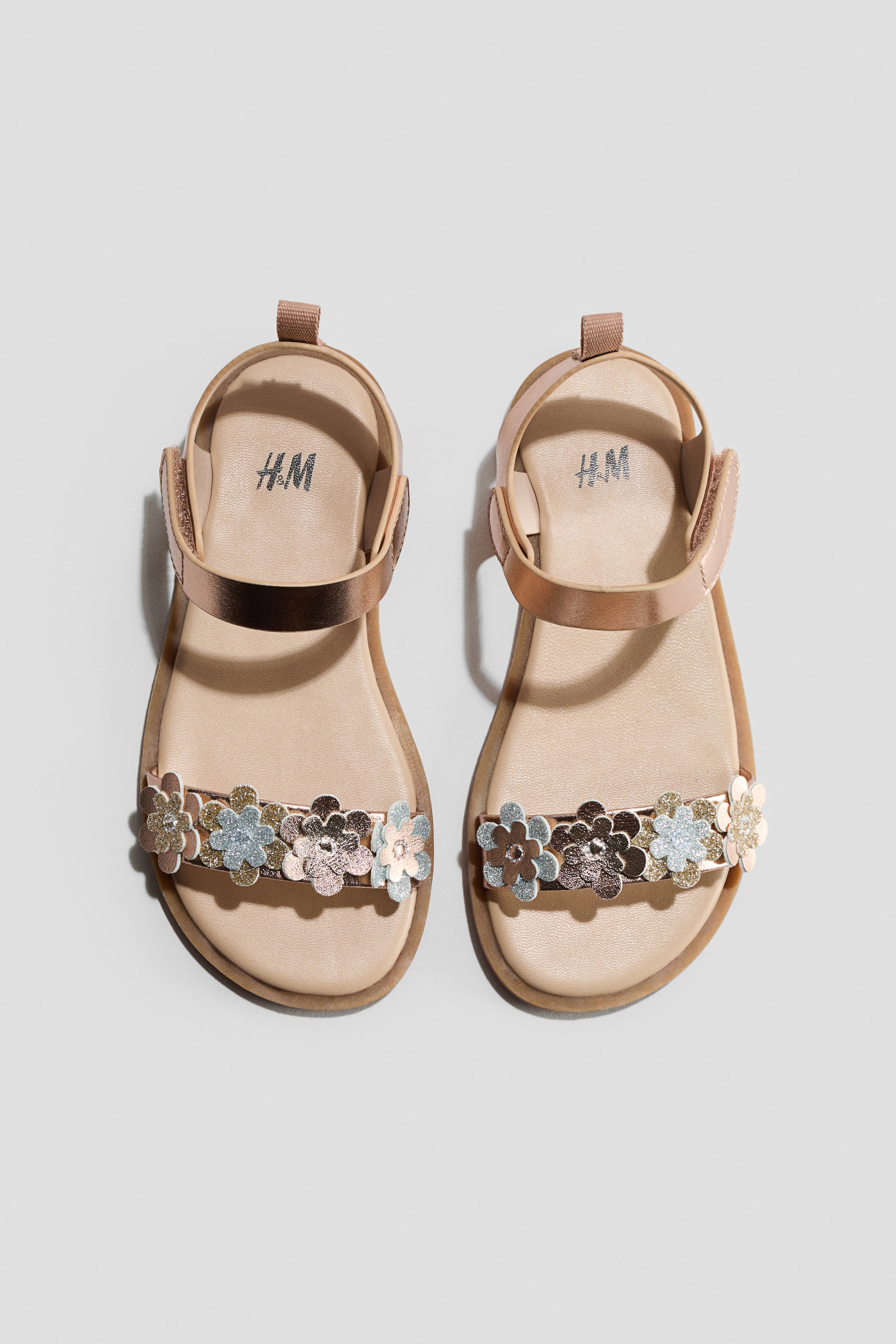 View larger image: A pair of children's sandals is lying flat, featuring a rose gold metallic finish and a light beige footbed displaying the H&M logo. Each sandal showcases a wide toe strap adorned with metallic flower appliques in rose gold, silver, and light blue, some with glitter, while an ankle strap secures the foot.
