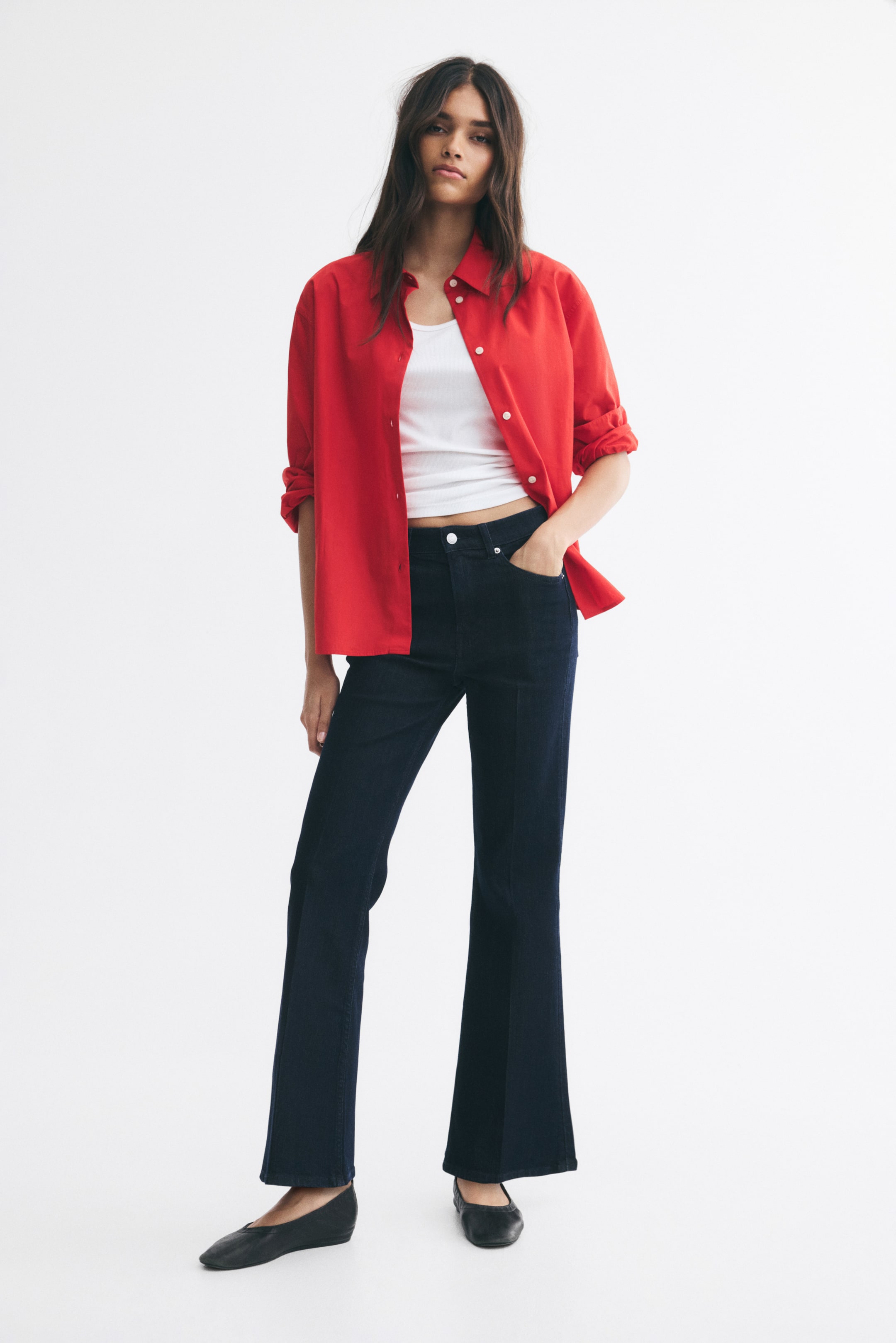 View larger image: High-waisted, dark-wash flared jeans featuring front creases are being worn by the model, who is standing with one hand in a pocket. The slim-fit denim jeans are styled with a white cropped tank top, an open red button-up shirt, and black ballet flats.