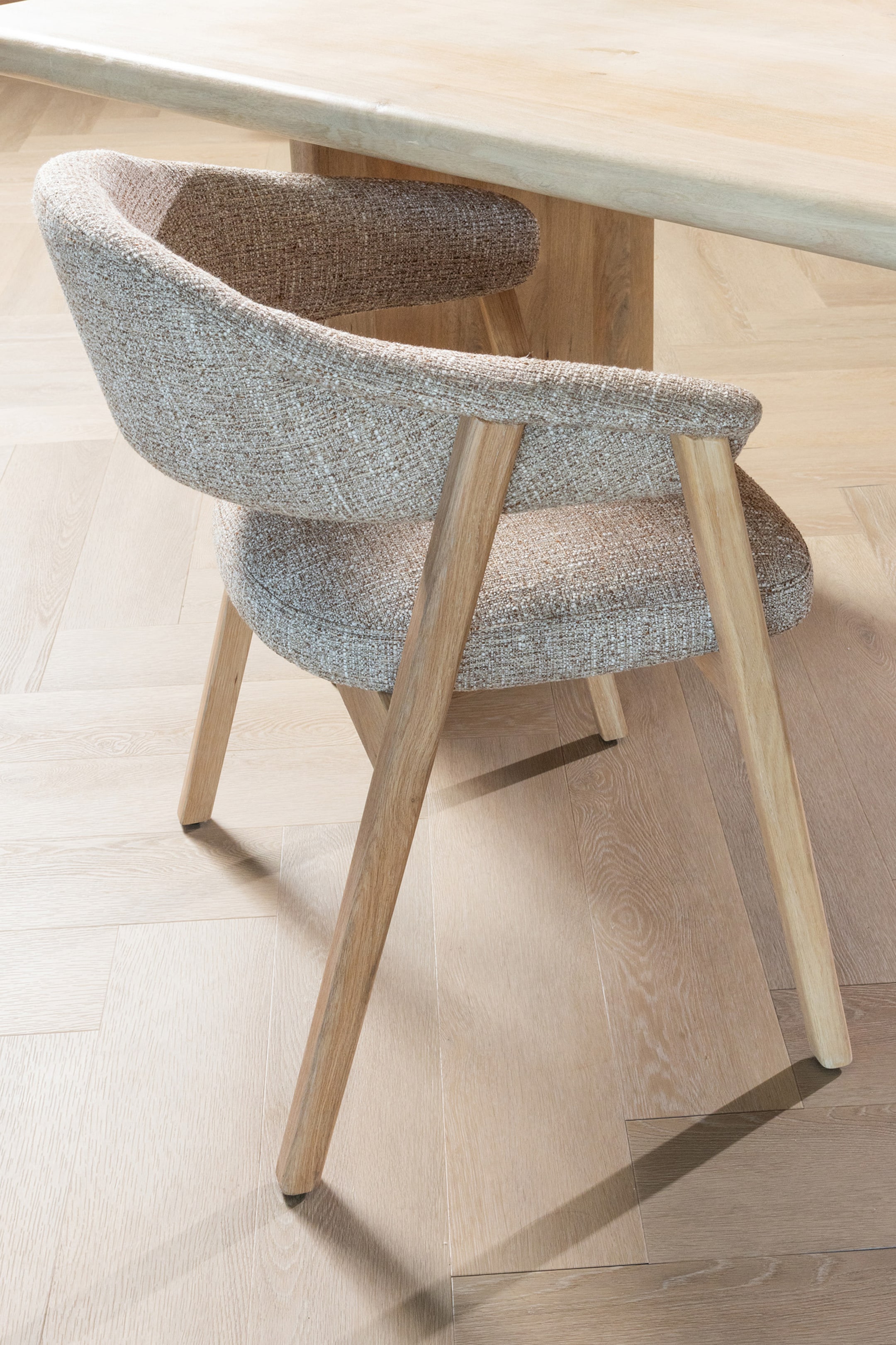 Agrandir l'image: Une chaise est recouverte d'un tissu moucheté brun clair, formant un dossier incurvé et des accoudoirs intégrés. Elle est présentée sous un angle arrière, mettant en valeur ses quatre pieds inclinés en bois clair.