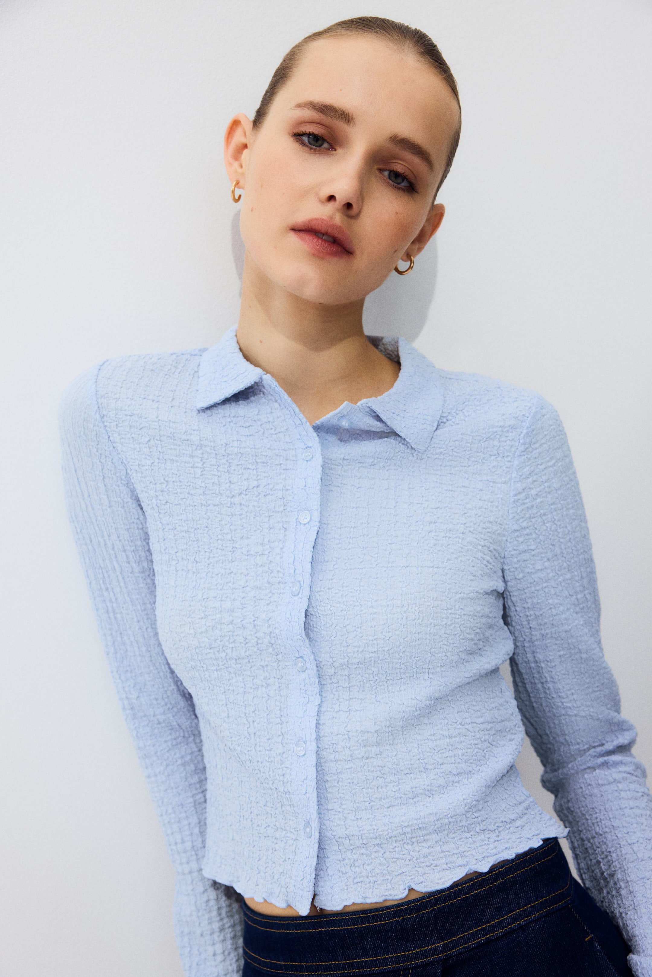 View larger image: A light blue long-sleeved shirt showcasing a distinctive puckered texture, a classic collar, and a button-front closure. Its scalloped bottom hem fits closely and is styled over dark denim.