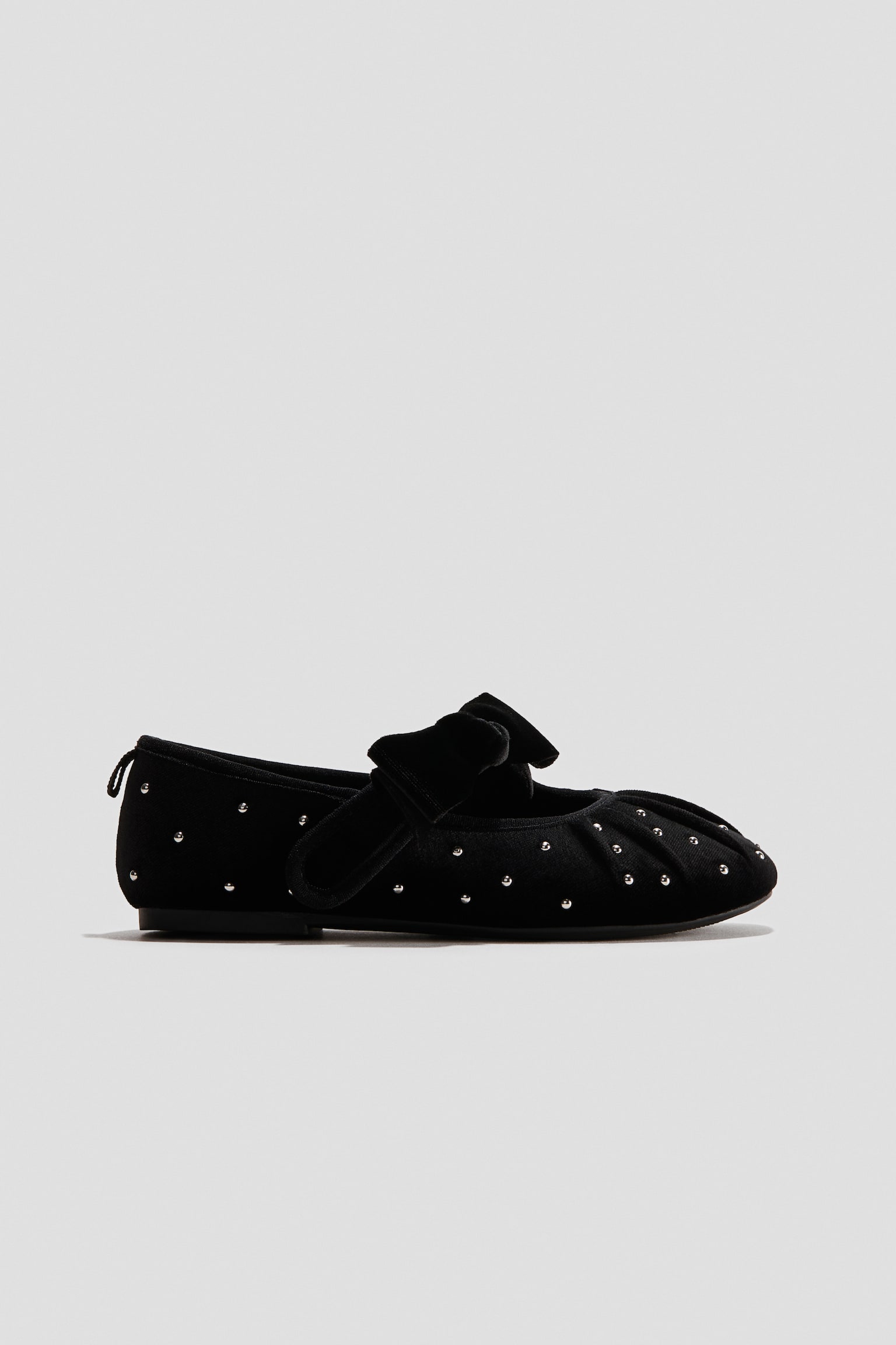Se muestra un zapato plano tipo ballet de terciopelo negro en vista de perfil lateral, con pequeños remaches plateados distribuidos sobre la parte superior y un sutil detalle fruncido en la punta redondeada. El zapato está diseñado con un moño decorativo de terciopelo a juego sobre el empeine y una suela delgada y plana.