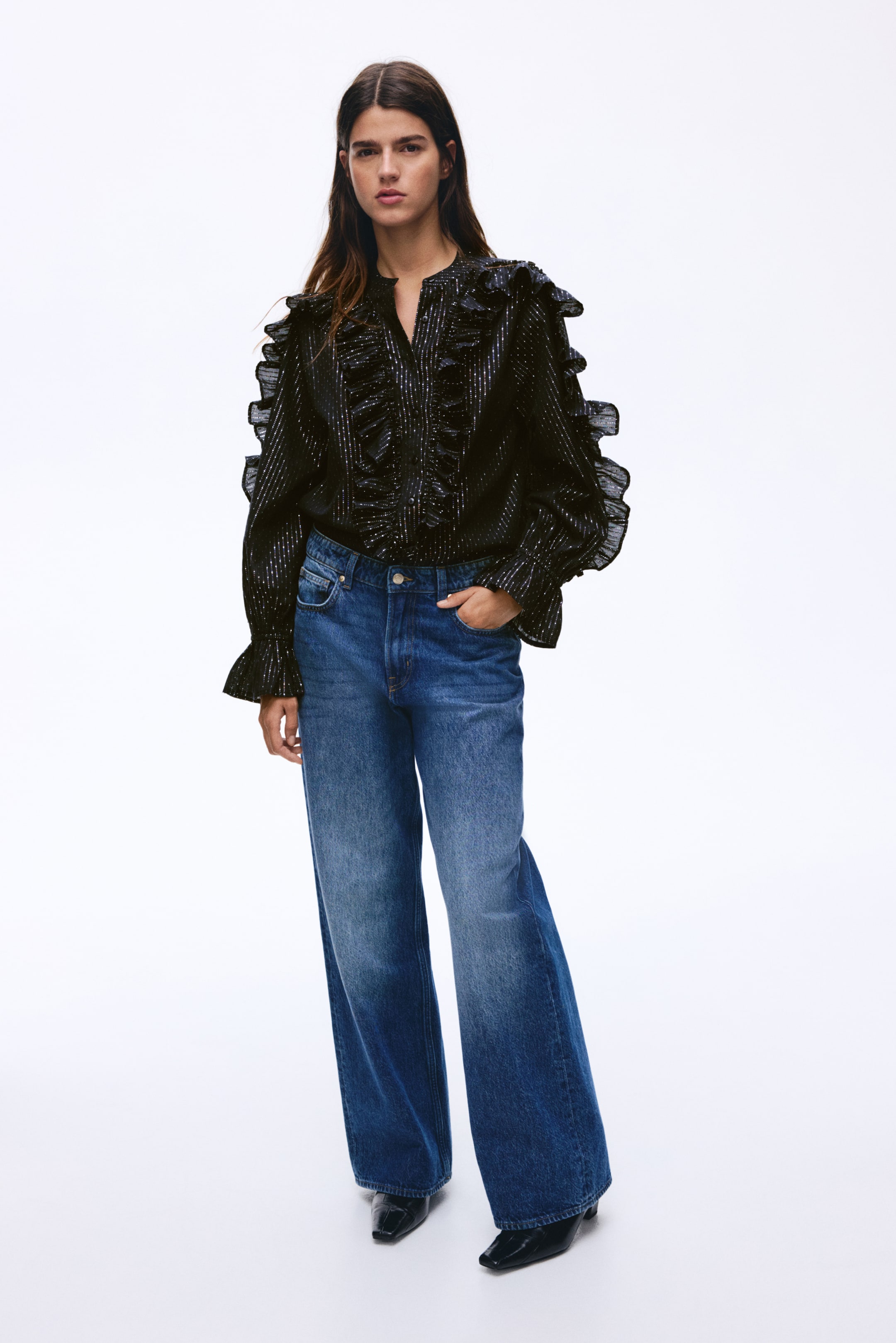 Agrandir l'image: Une blouse bleu foncé, ornée de rayures verticales scintillantes, avec un col officier et un décolleté en V, ainsi que des volants en cascade sur les épaules, le devant et les poignets, est portée rentrée dans un jean bleu.