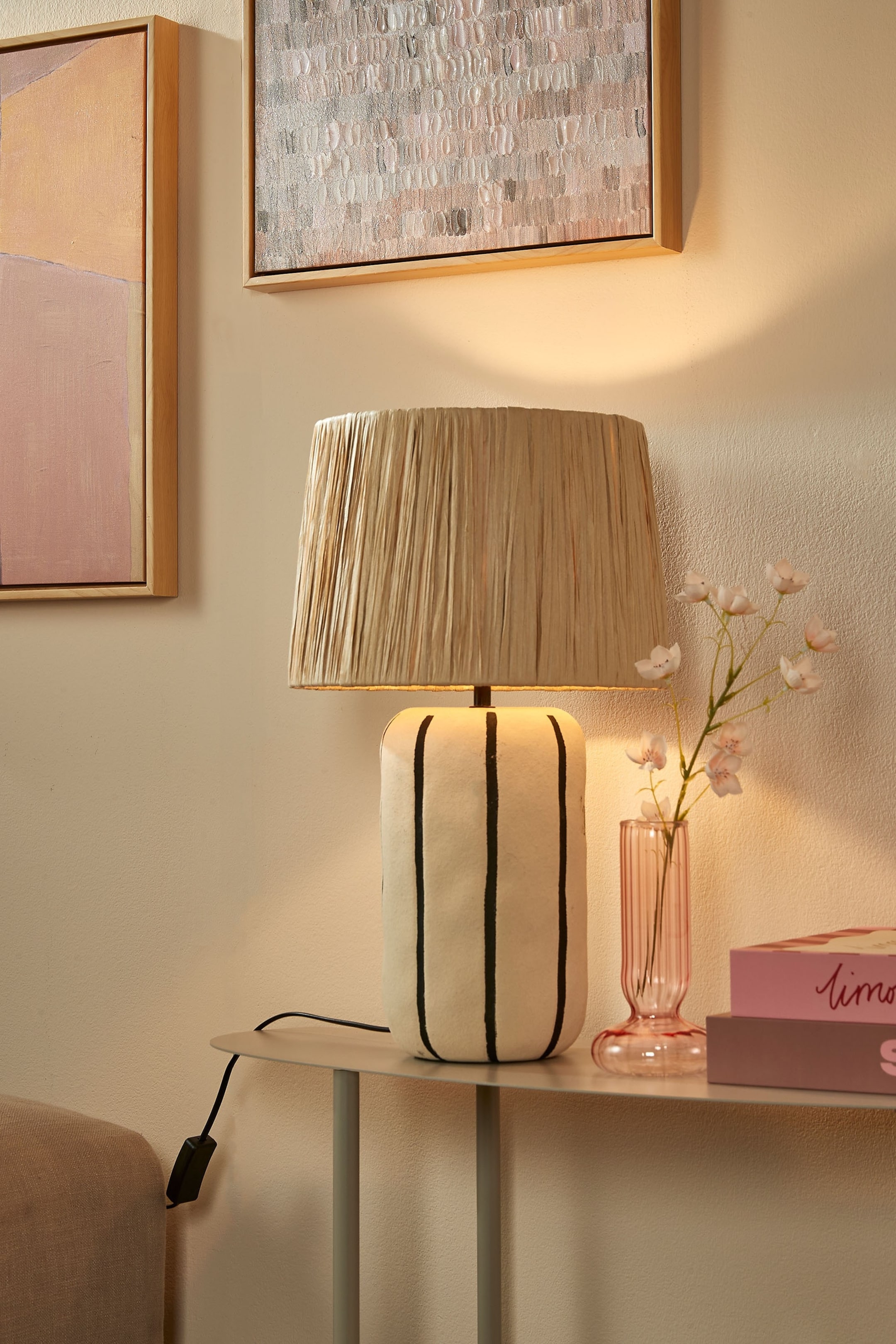 Agrandir l'image: Une lampe de table présente un abat-jour évasé et plissé, fabriqué à partir de fibres aux tons naturels, et un socle cylindrique blanc cassé orné de rayures verticales noires irrégulières. La lampe est allumée, positionnée face à l'avant.