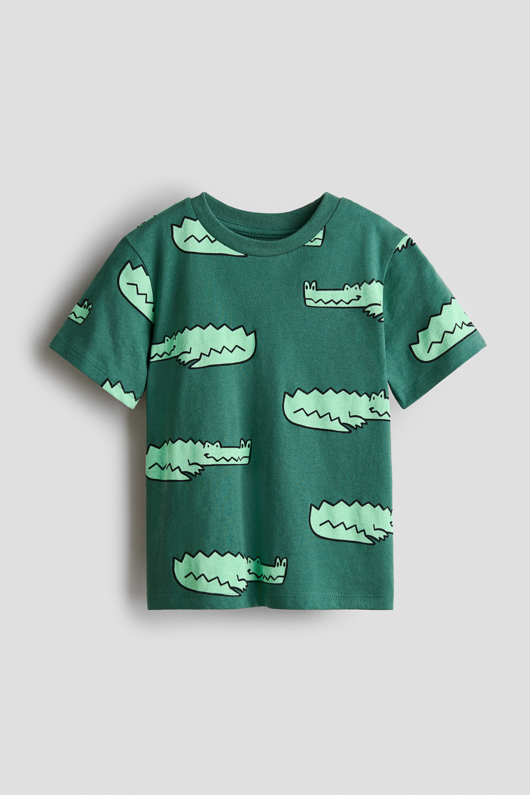拡大画像を表示: プリントコットンジャージーのダークグリーンTシャツが前面に表示されています。丸みを帯びたリブトリムのネックライン、優しく落ちた肩、そしてライトグリーンのカートゥーンワニのパターンが特徴です。