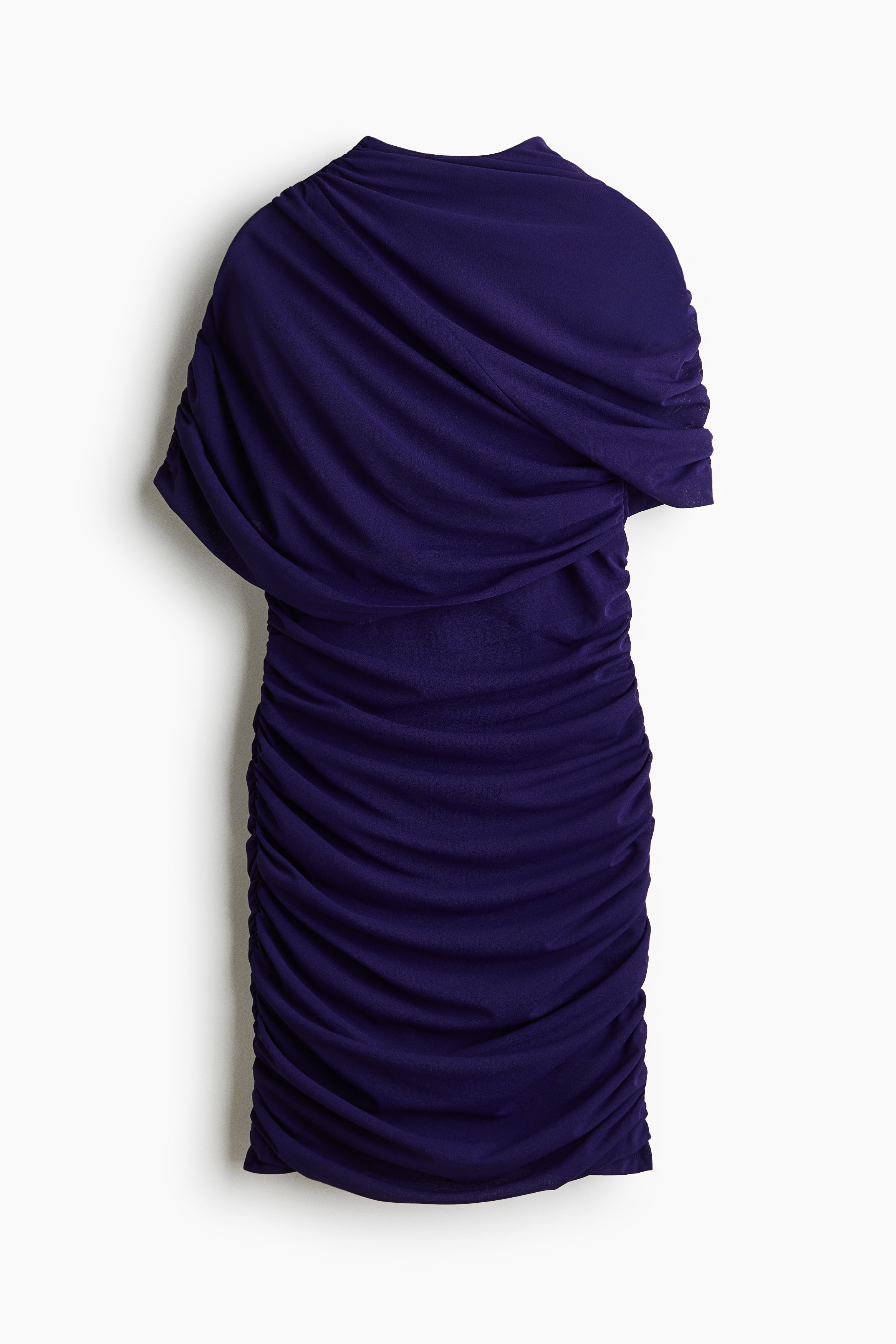 A dark purple fitted mini dress featuring extensive ruching and a draped, cape-like layer across the shoulders is worn by the model. The dress is designed with a high neckline and a single short sleeve visible on one side.