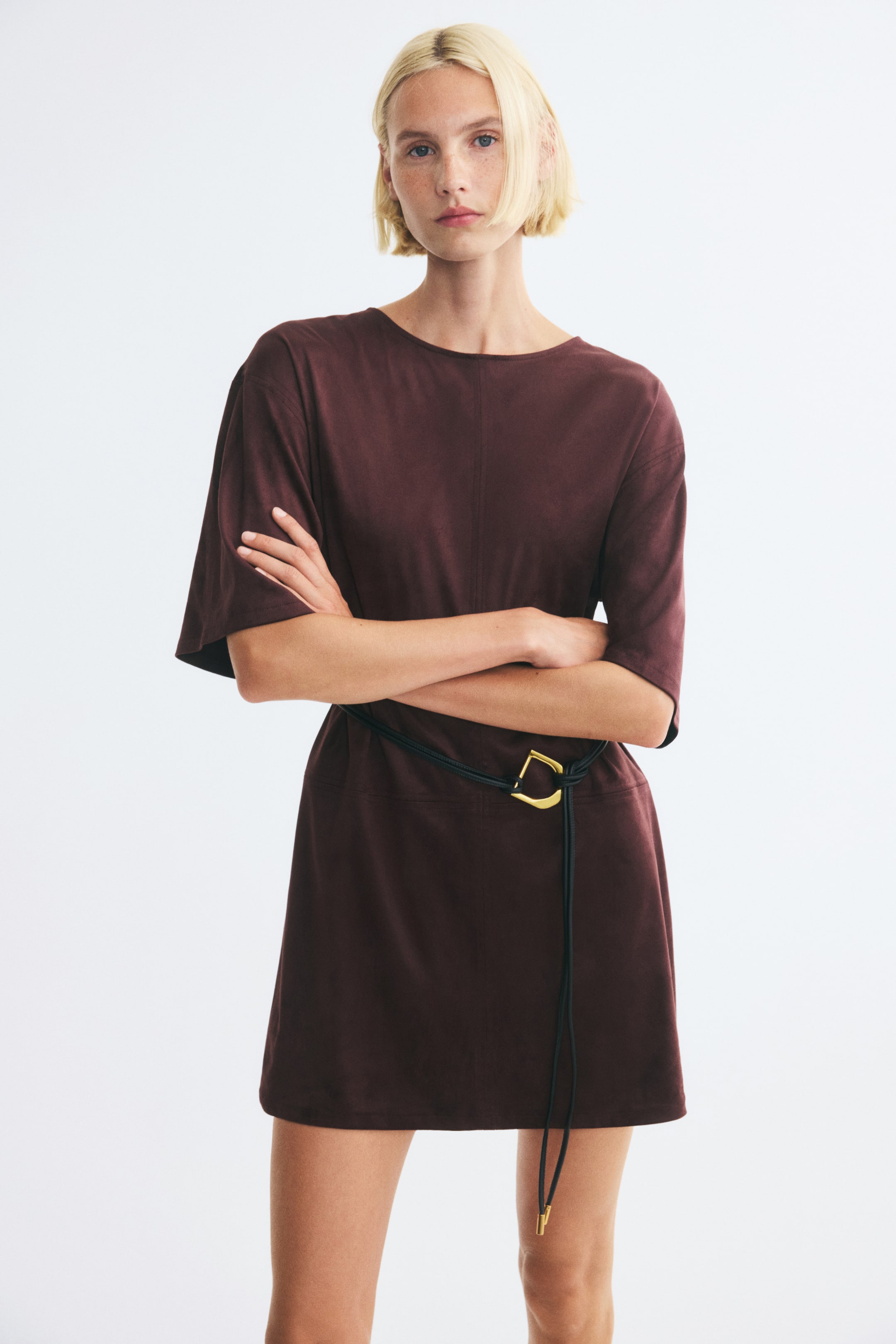 Voir une image plus grande: Une robe courte à coupe ample en tissu duveteux bordeaux foncé est portée par le mannequin, avec un col rond, des épaules tombantes et une couture centrale visible à l’avant. La robe est cintrée à la taille avec une fine ceinture à cordon noire munie d’une boucle couleur or, et le mannequin est debout, les bras croisés.