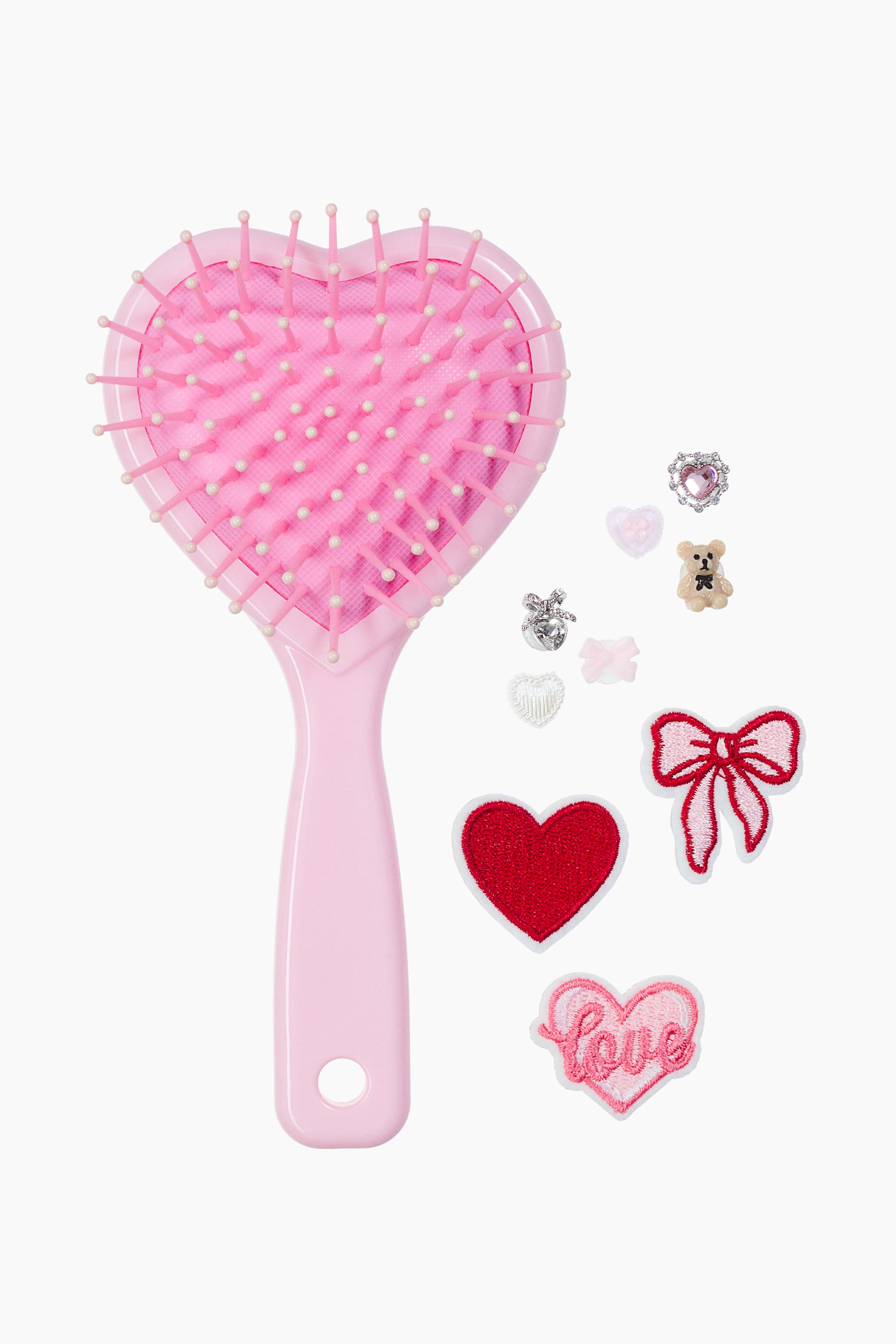 Visualizza immagine più grande: Una spazzola per capelli rosa chiaro è rivolta verso la parte anteriore, mettendo in mostra una testina a forma di cuore con setole rosa con puntine arrotondate e un manico liscio con un foro circolare. Accanto alla spazzola sono presenti diversi piccoli elementi decorativi, tra cui toppe ricamate e charm in plastica con design a cuore, fiocco e orsetto.