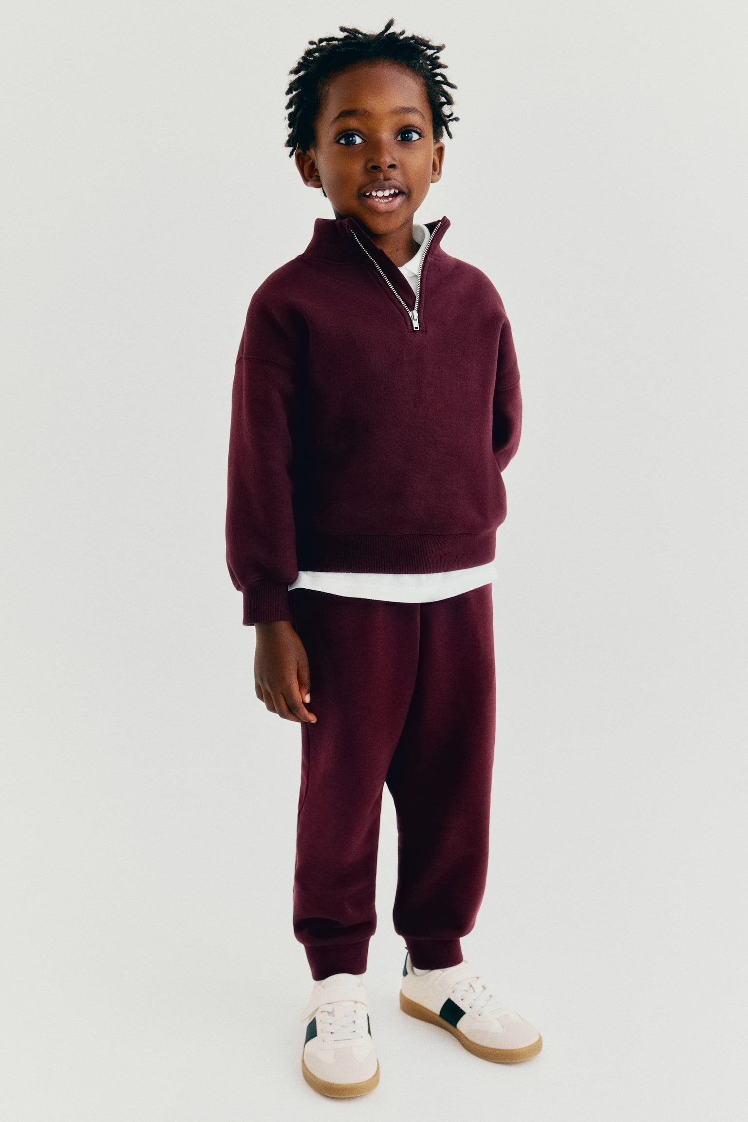 Burgundy track pants in sweat fabric are shown in a relaxed fit with ribbed cuffs at the ankles, viewed from the front. They are paired with a matching half-zip sweatshirt to complete the coordinated look. - 1