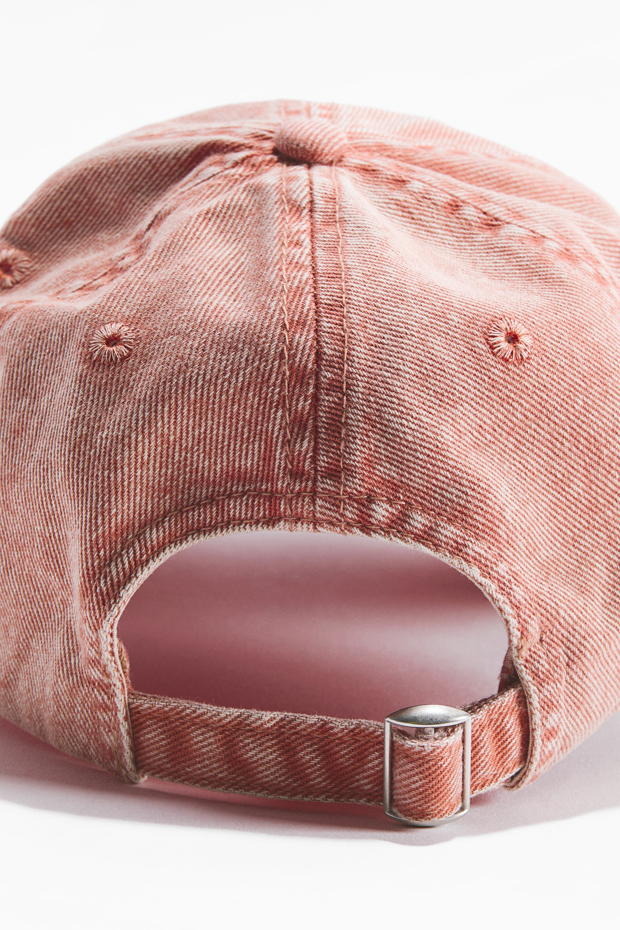 View larger image: A washed reddish-orange cap, crafted from cotton twill, is displayed from the rear. It boasts a fabric tab with a silver-toned metal slide buckle at the back for adjustment, complemented by visible stitching on its panels and embroidered eyelets.