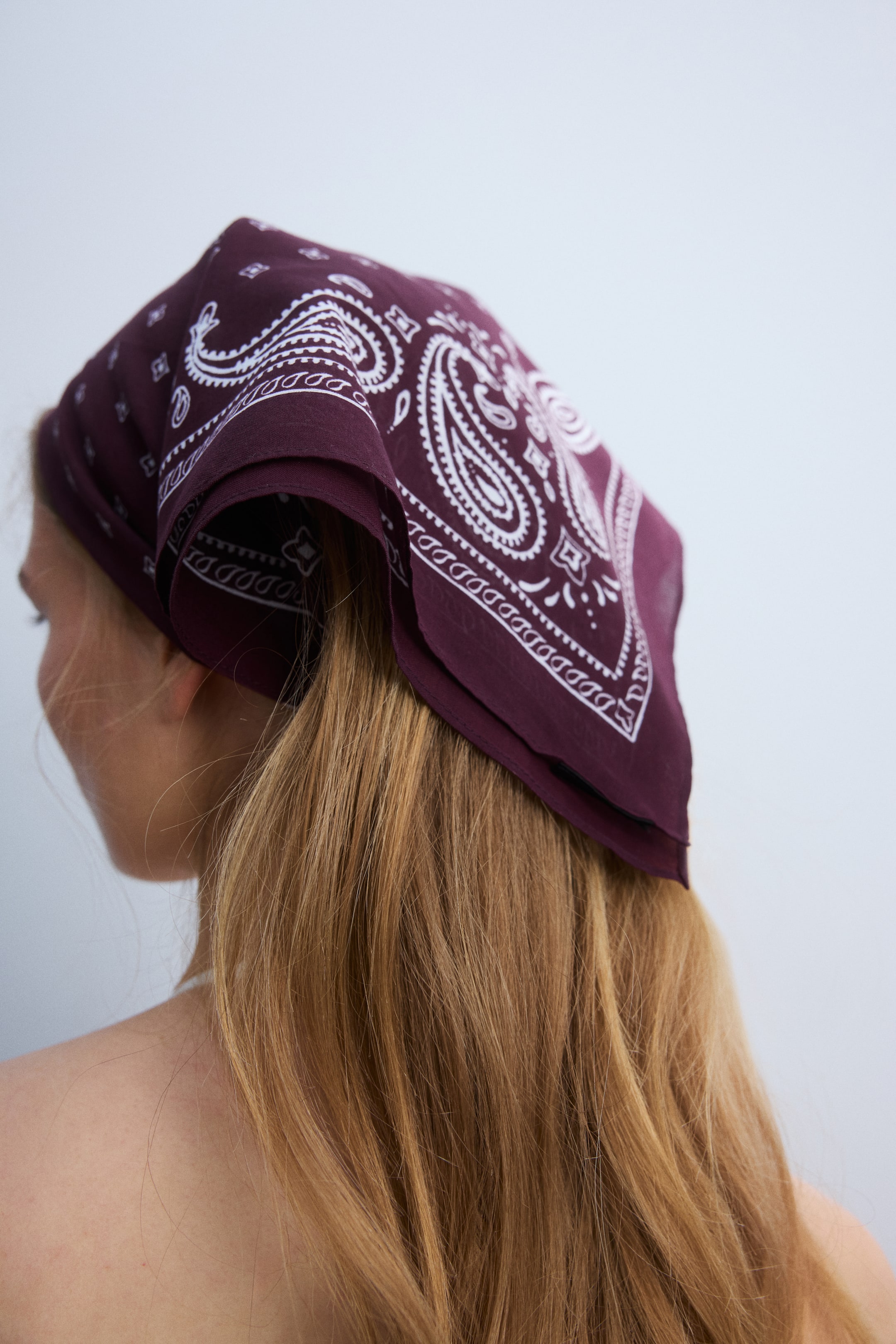 Zobrazit větší obrázek: Burgundský šátek s bílým paisley vzorem je uvázán na zadní straně hlavy, jeho složená látka spočívá na světle hnědých vlasech.