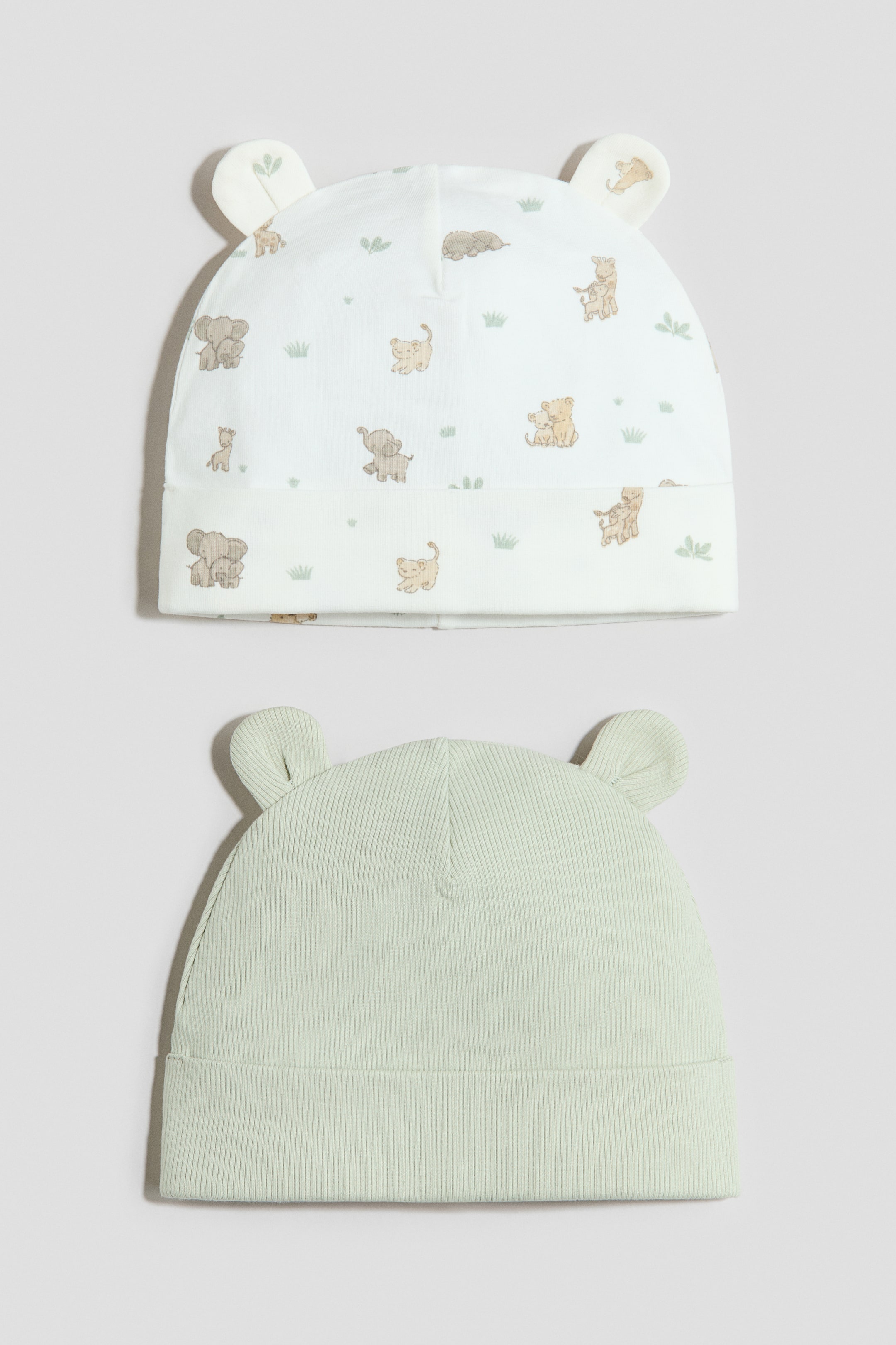 View larger image: A pack of 2 ear-detail beanies is presented, both featuring folded brims and shown facing forward. One beanie is white with a repeating animal print, and the other is light green with an all-over ribbed texture.