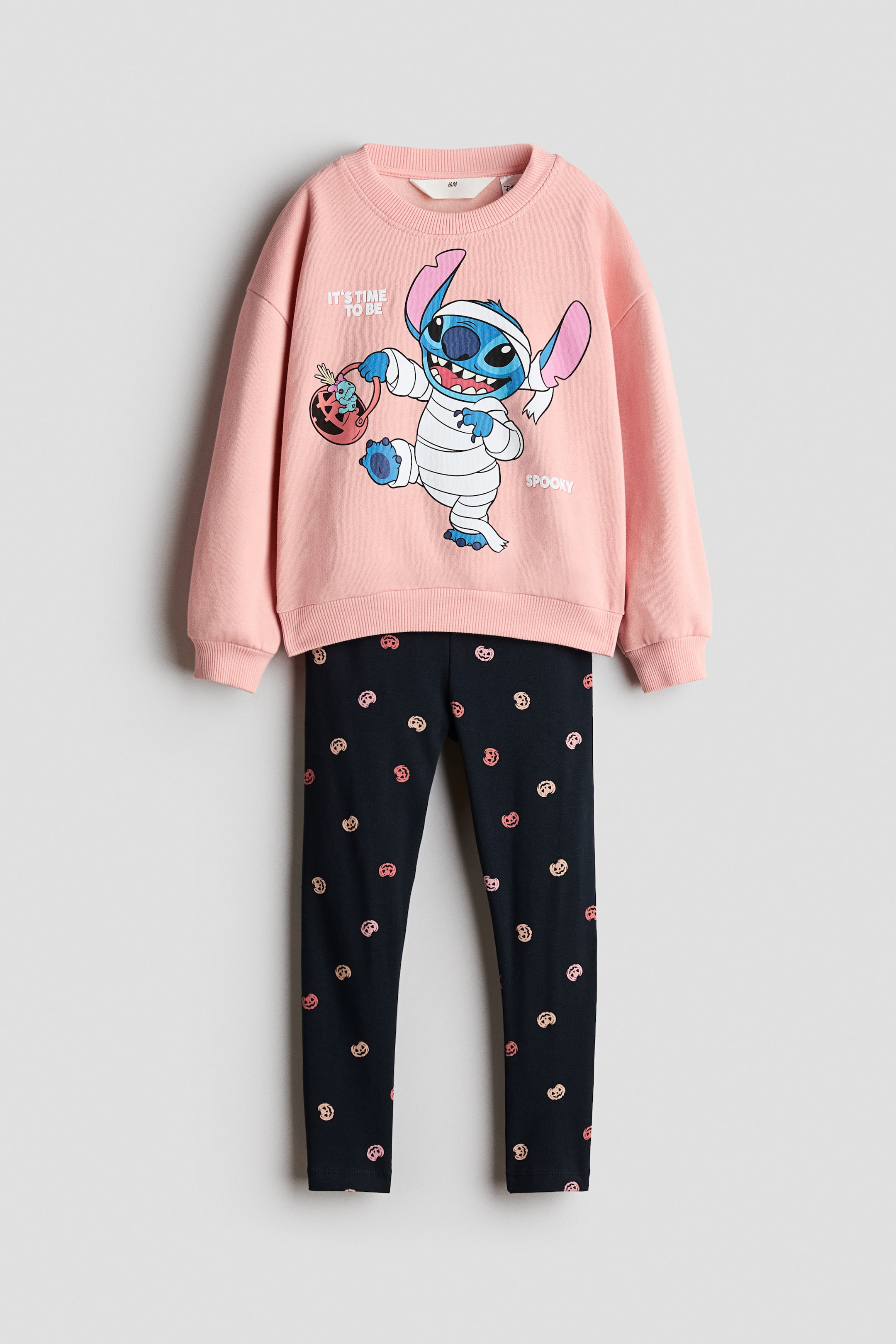 Visualizza immagine più grande: Questo completo a due pezzi, visto frontalmente, presenta un maglione rosa chiaro a maniche lunghe e leggings neri. Il maglione presenta un girocollo rifinito a coste, spalle scese e una grafica di Stitch vestito da mummia, mentre i leggings coordinati mostrano una stampa all-over di piccole zucche rosa.