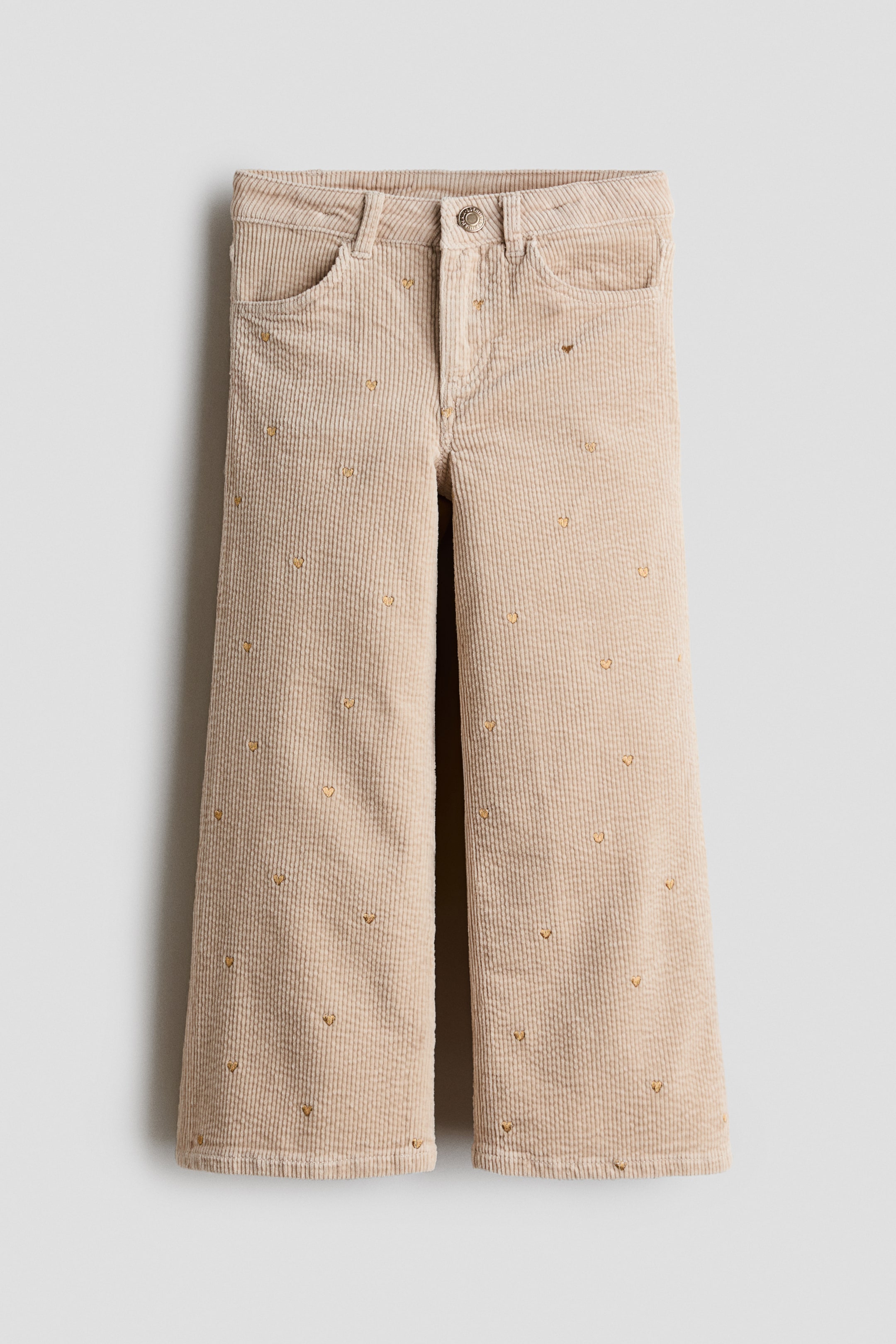 Voir une image plus grande: Une paire de pantalons en velours côtelé beige est présentée de face, avec une coupe à jambe large ornée d’un motif dispersé de petits cœurs dorés brodés. Le pantalon affiche un design classique à cinq poches, avec une ceinture, des passants de ceinture et une fermeture à bouton sur le devant.