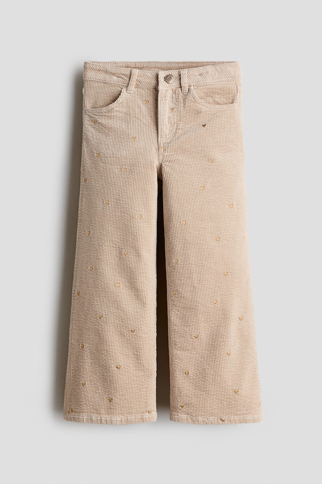 A pair of beige corduroy trousers is facing front, featuring a wide-leg cut decorated with a scattered pattern of small, embroidered gold hearts. The trousers are showing a classic five-pocket design with a waistband, belt loops, and a button-front closure. - 1