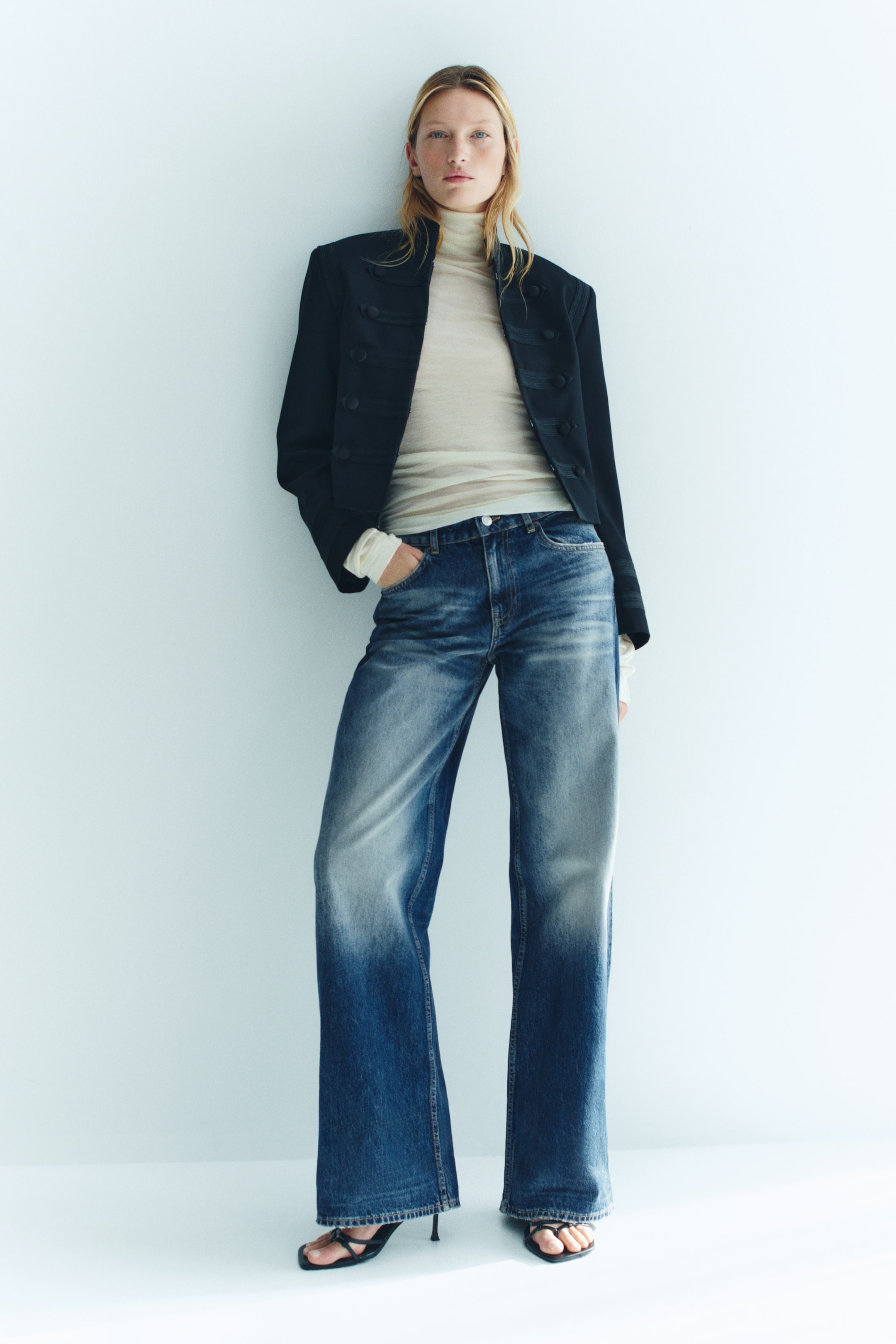 Visualizza immagine più grande: Jeans in denim blu a gamba ampia con lavaggio slavato, indossati dalla modella che è in piedi con una mano appoggiata nella tasca anteriore. I jeans a vita regolare sono abbinati a un top dolcevita trasparente color crema, a una giacca scura strutturata e a sandali neri con tacco.