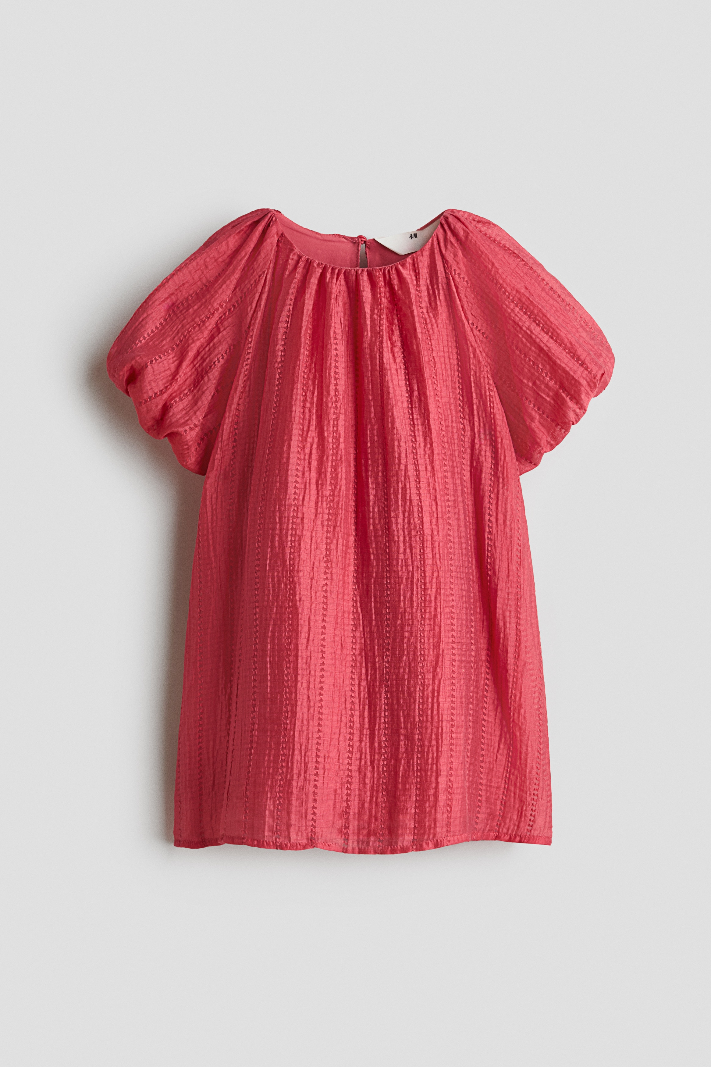 A red A-line dress in a soft, textured weave is shown from the front, featuring a gathered round neckline and short, voluminous raglan sleeves with elasticated cuffs. A red, heart-shaped crossbody bag is draped over the front of the dressâs loose silhouette.